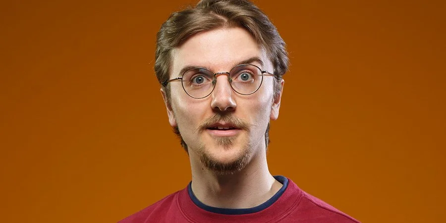 Comedian Dru Cripps with brown hair, glasses, a goatee, wearing a red shirt, against an orange background.