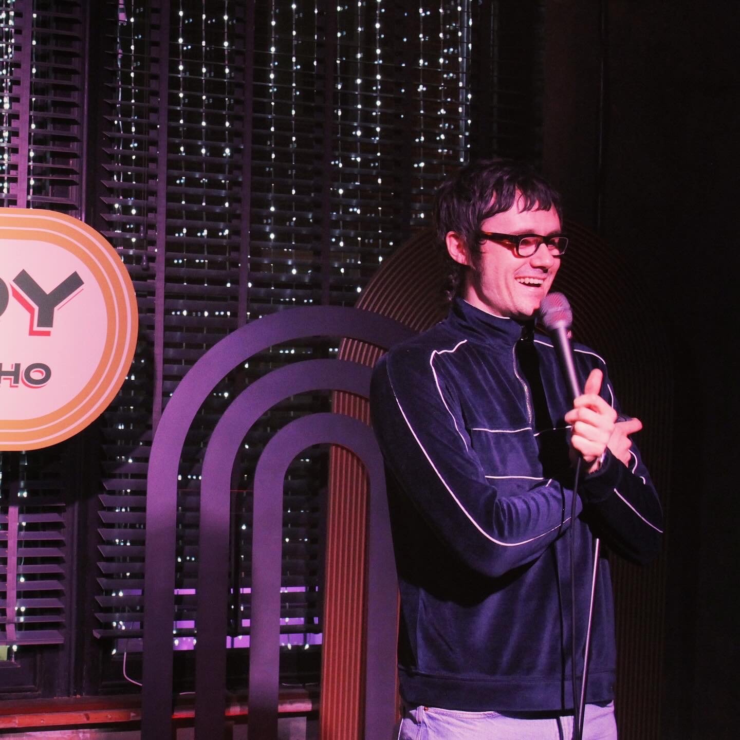 Comedian Bert Broadbent on stage at Comedy Club Comedy Union in Soho with glasses and dark hair is holding a microphone and smiling on stage with purple-lit background.