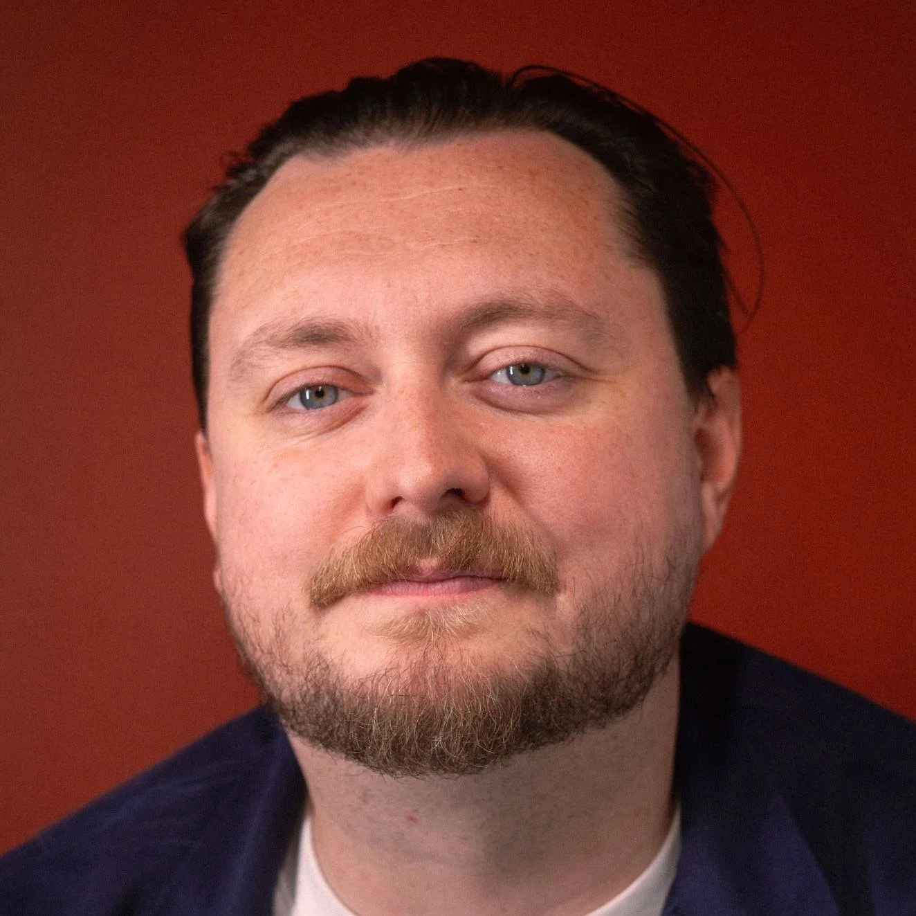 Comedian Eli HArt with light skin, blue eyes, a reddish-blonde beard, and mustache, against a maroon background.