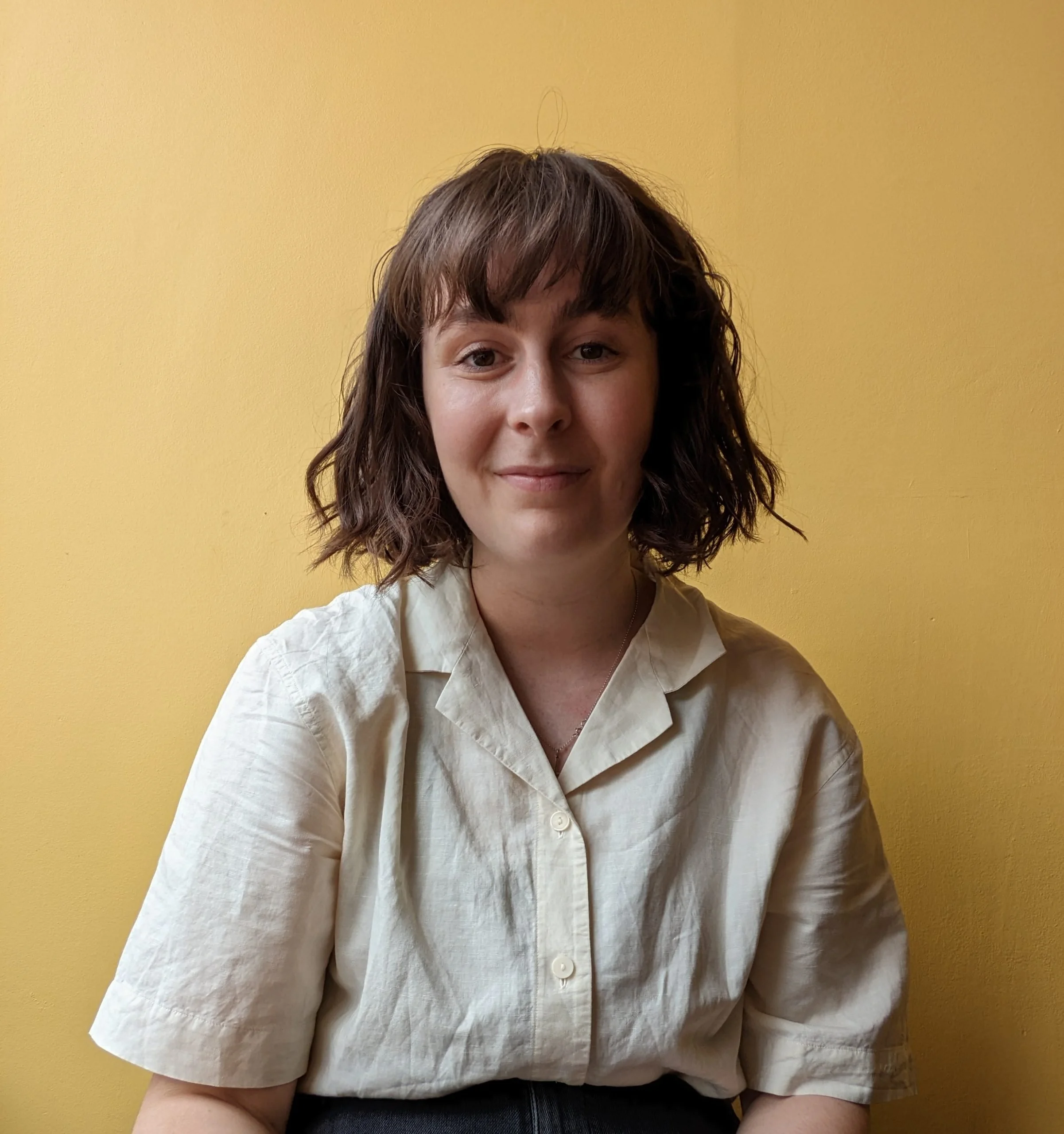 Comedian Laura Walsh with shoulder-length wavy brown hair and fair skin, wearing a white button-up shirt, standing in front of a yellow wall, smiling slightly.