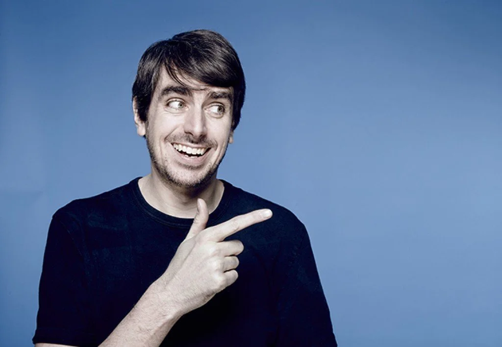 Comedian Darren Walsh with brown hair, wearing a black shirt, smiling and pointing to his right against a blue background.