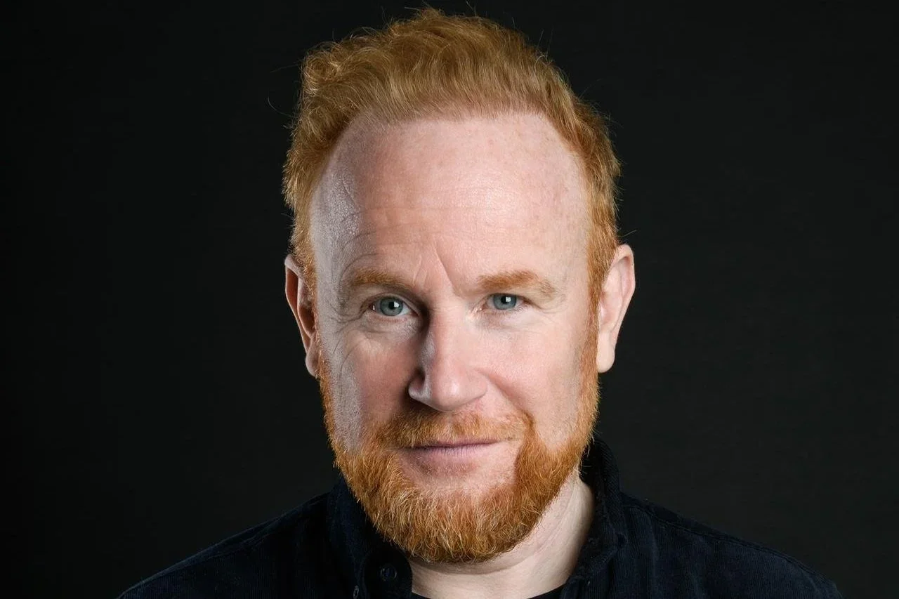 Comedian Rory O'Hanlon with a beard, wearing a black shirt, against a black background.