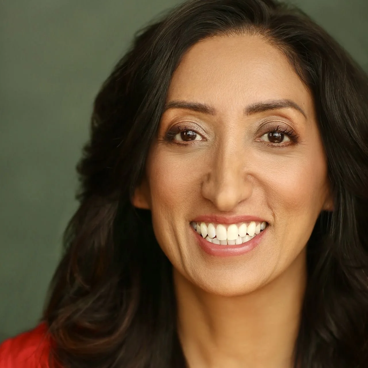 Comedian Shazia Mirza smiling woman with dark hair and brown eyes, wearing makeup and a red top against a green background.