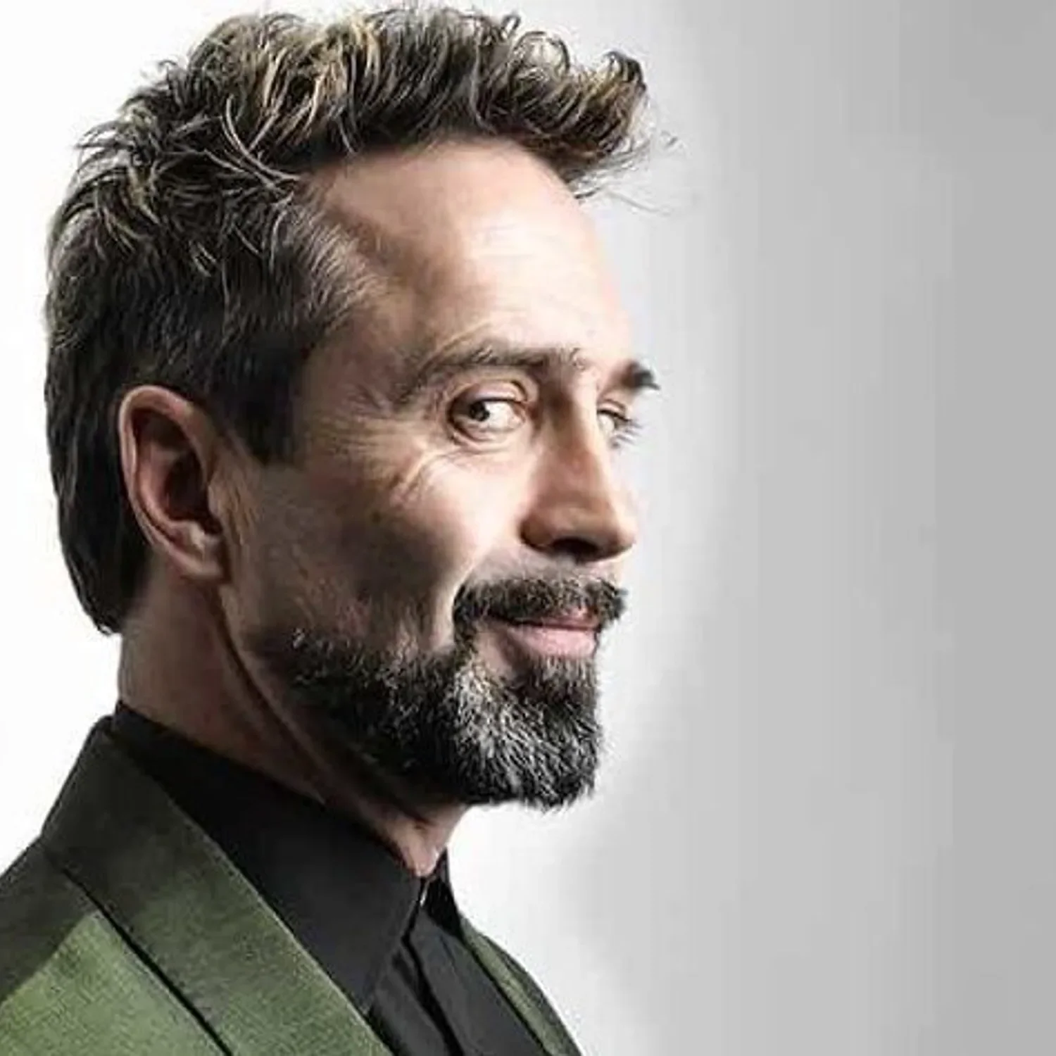 Comedian Phil Nichol with a beard and mustache, looking slightly to the right, wearing a dark shirt and a green jacket, against a plain light background.