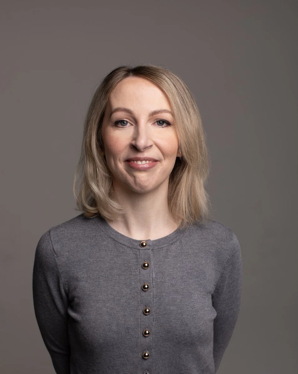 Comedian Jenny Collier with blonde hair wearing a gray sweater, smiling slightly, against a neutral gray background.