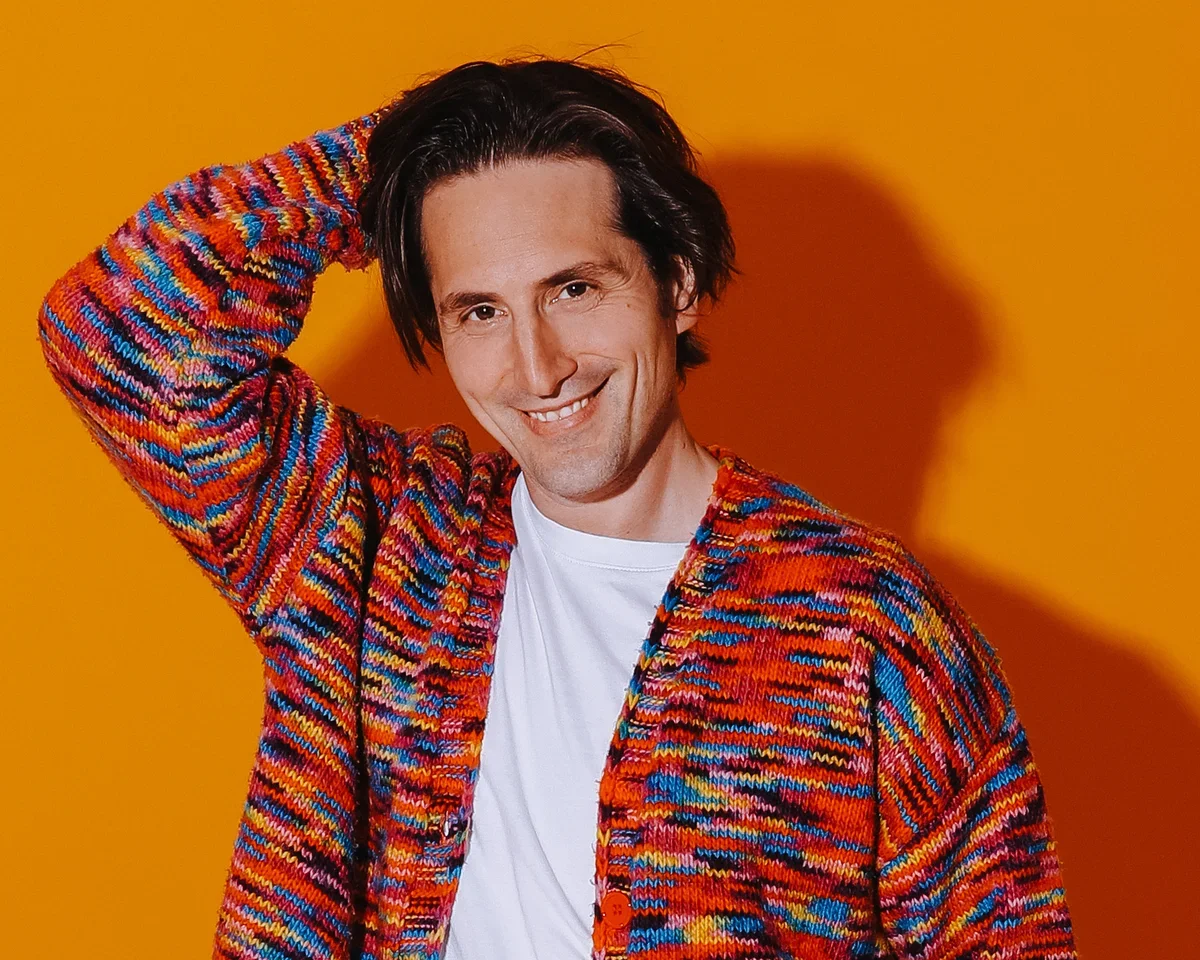 Comedian Luke McQueen  smiling, wearing a colorful patterned sweater and a white t-shirt, standing against an orange background.