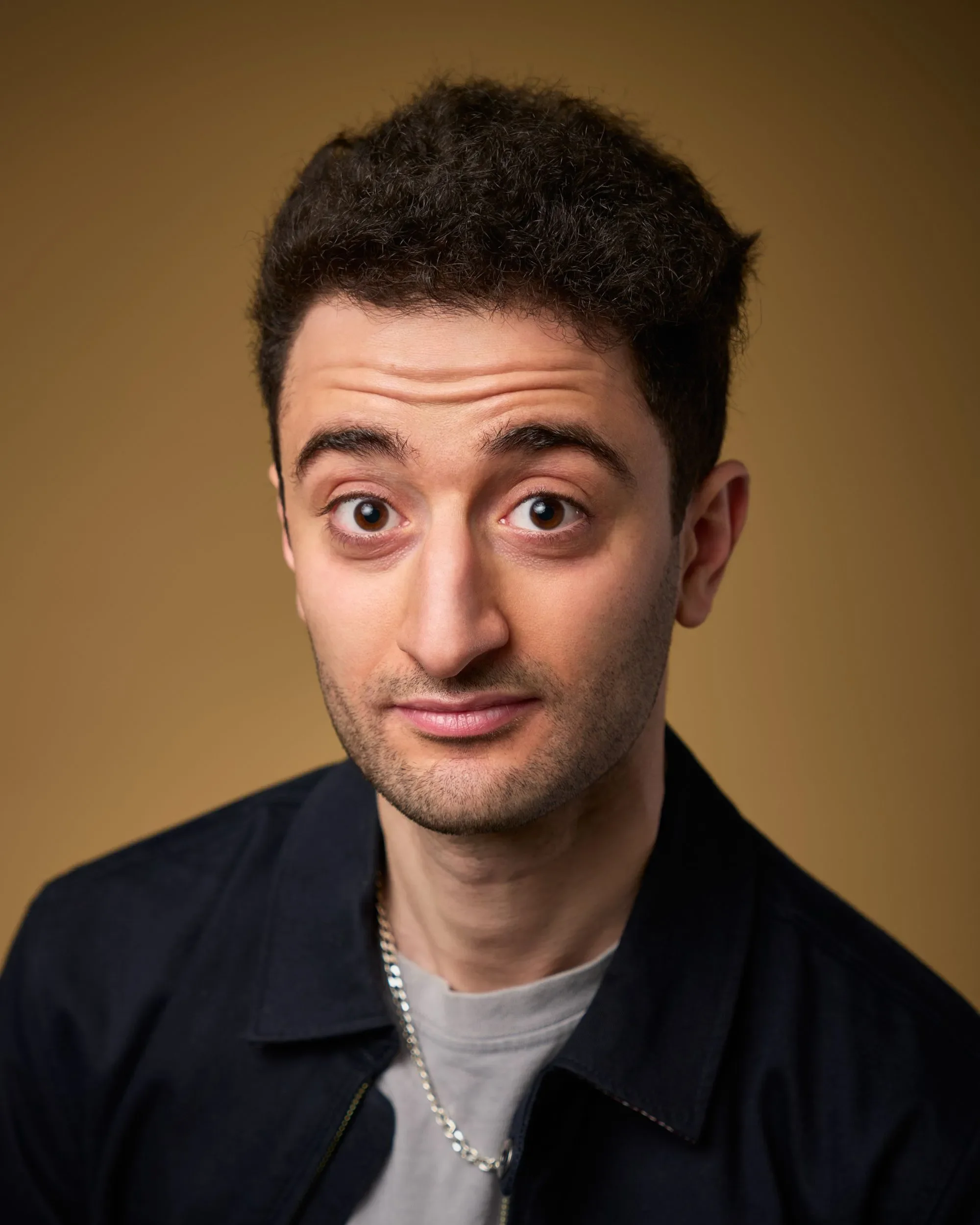 Comedian Hasan Al-Habib dark, curly hair and brown eyes is fading out, showing only his eyes, forehead, nose, mouth, and part of his neck. He is wearing a black jacket, a gray shirt, and a silver chain necklace against a plain brown background.