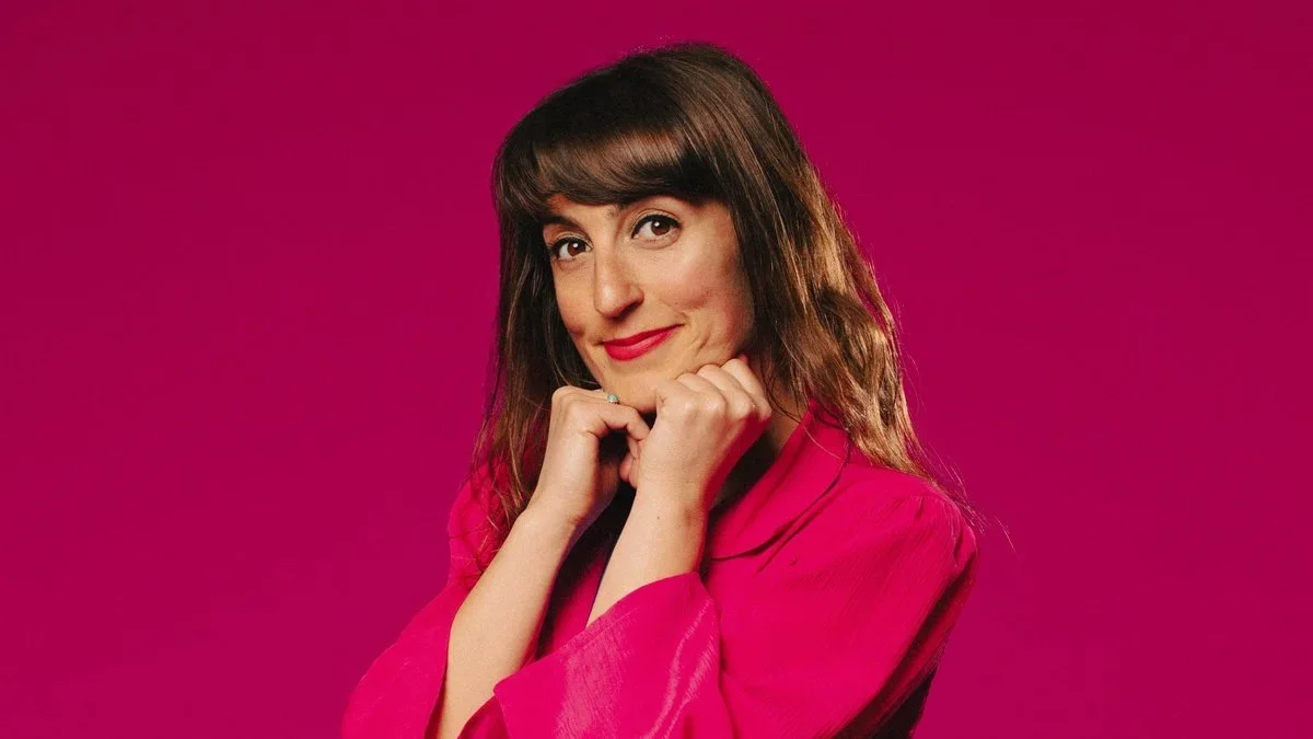 Comedian Jo Griffin with medium-length brown hair and bangs, wearing a bright pink blazer, smiling softly with her chin resting on her hands against a matching pink background.