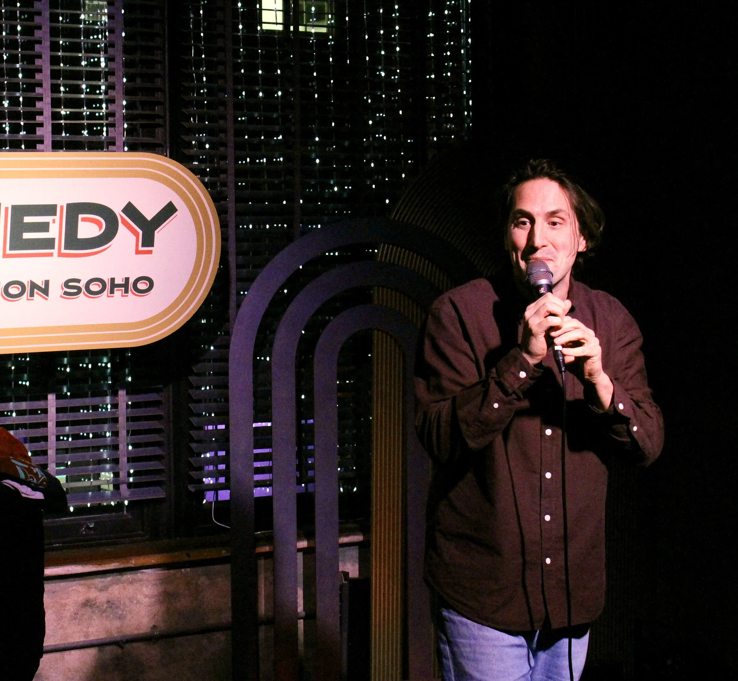Comedian Luke McQueen performing at Soho Comedy Club The Union Club with long dark hair holding a microphone, standing in front of a sign The background features a dark wall with a grid of small lights and a curved decorative element.