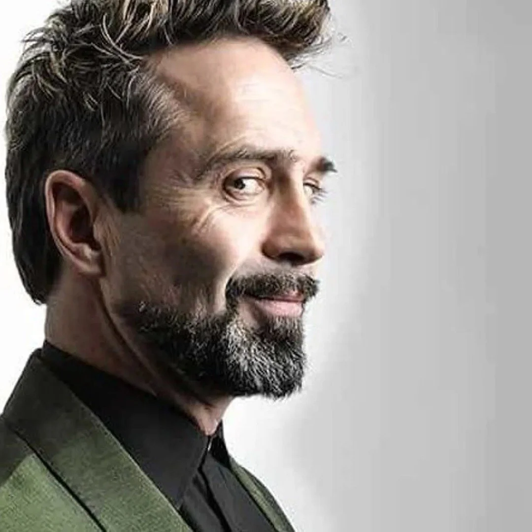 Comedian Phil Nichol with a beard and mustache, partly smiling, looking over his shoulder, with gray hair and dressed in a dark shirt and green jacket.