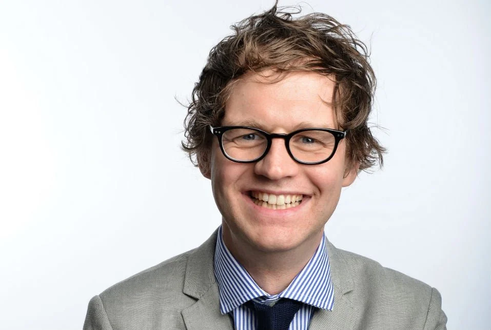 Comedian Mark Dolan with curly brown hair, black glasses, and a friendly expression. He's wearing a gray suit, a blue and white striped shirt, and a navy tie, standing against a plain white background.