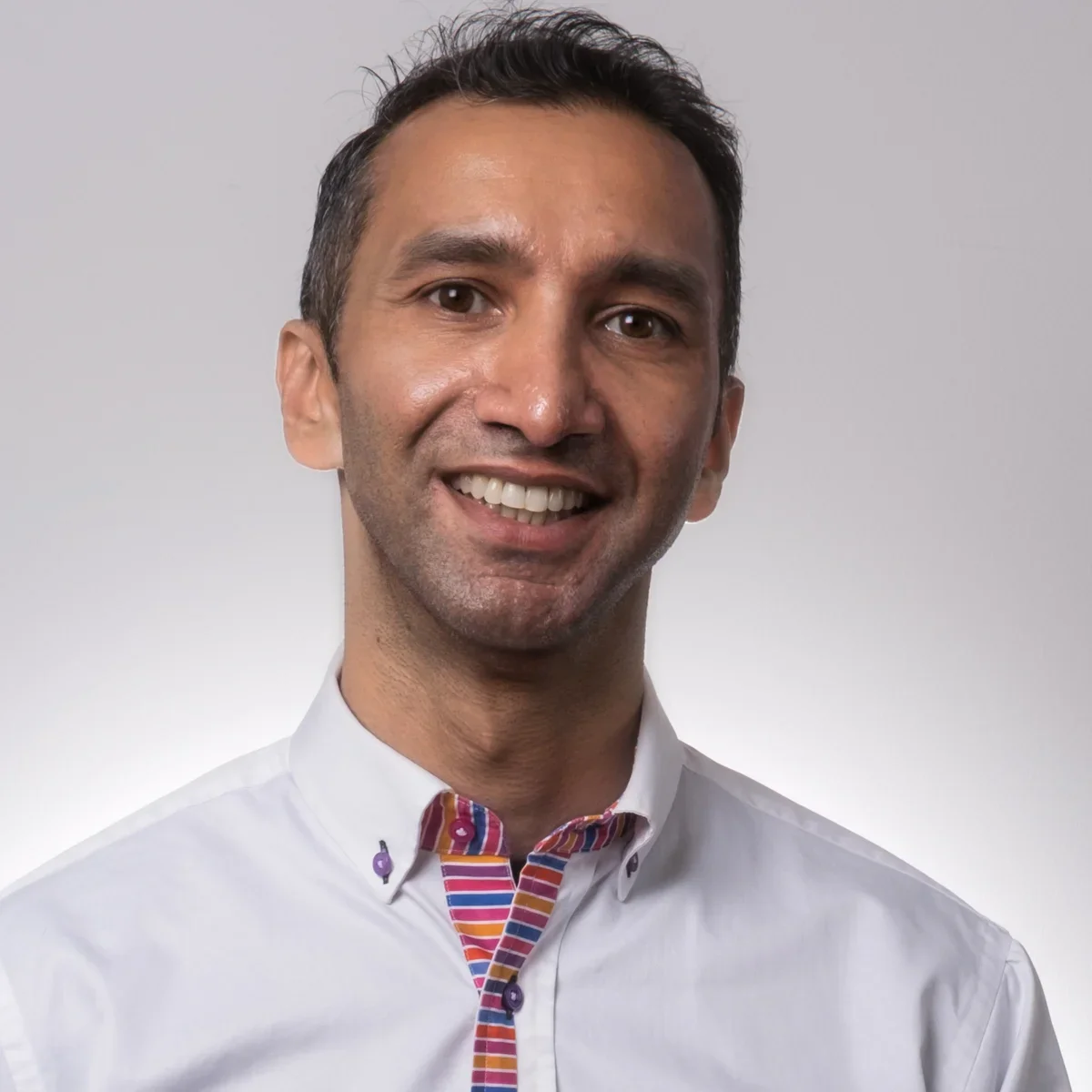 Comedian Imran Yusuf with dark hair, wearing a white shirt with a colorful striped collar and inner placket, against a light gray background.