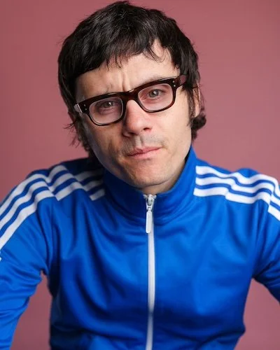 Comedian Bert Broadbent wearing glasses and a blue Adidas track jacket with white stripes against a pink background.