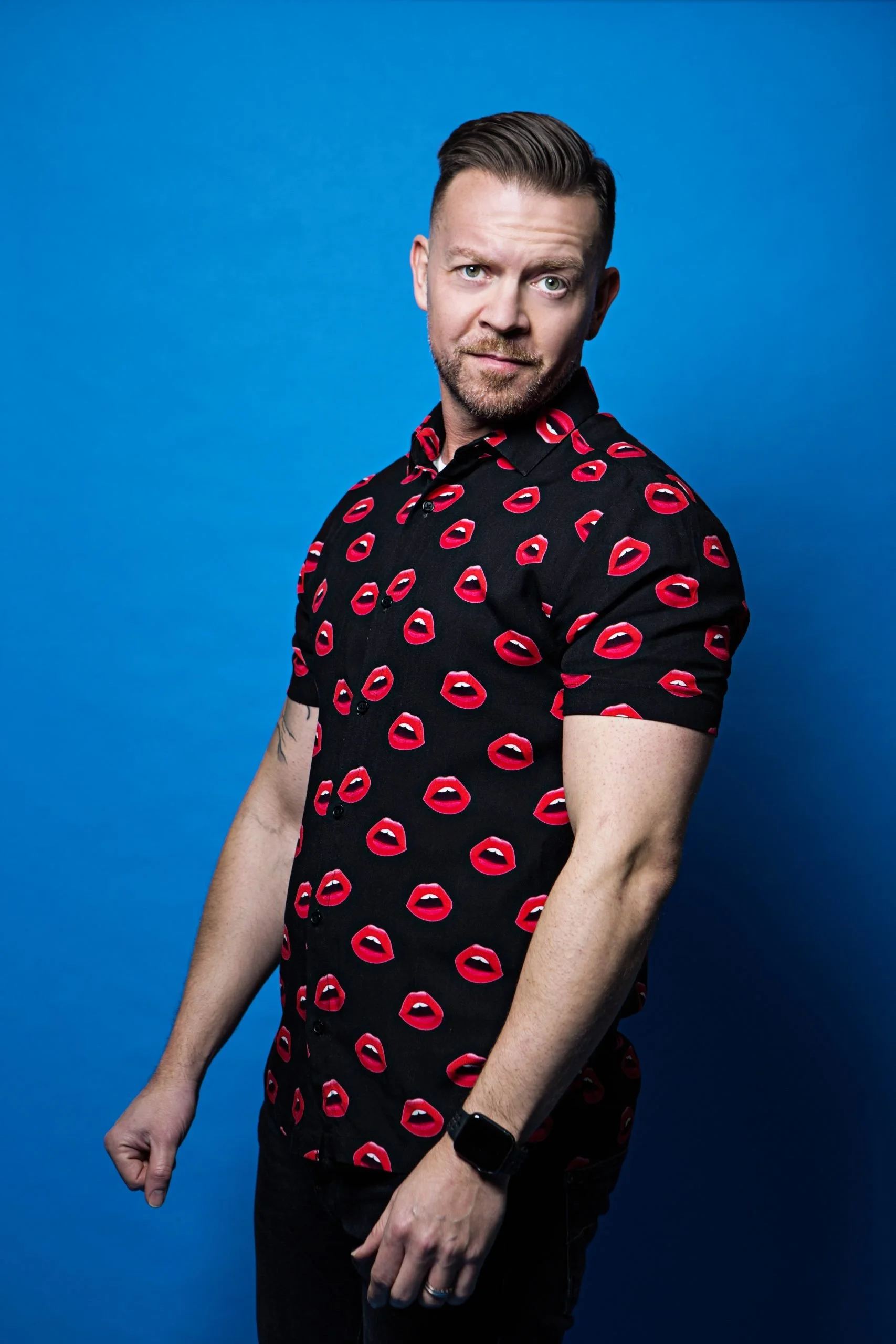 Comedian Russell Arathoon short, styled hair, a beard, wearing a black shirt with red lips pattern, standing against a solid blue background.