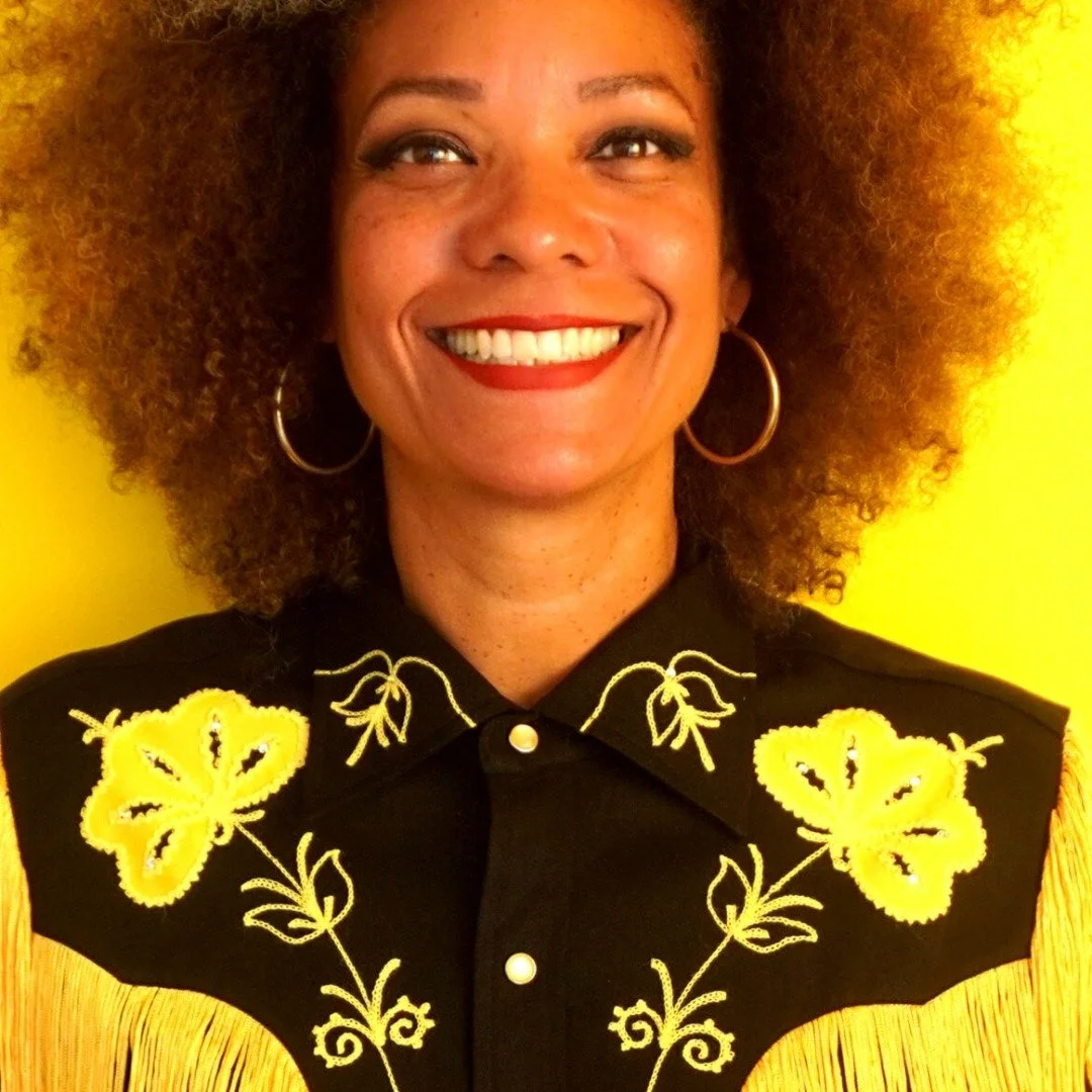 Comedian Malinda Mukuma with voluminous curly hair, wearing a black shirt with yellow floral embroidery and fringe details, smiling in front of a yellow background.