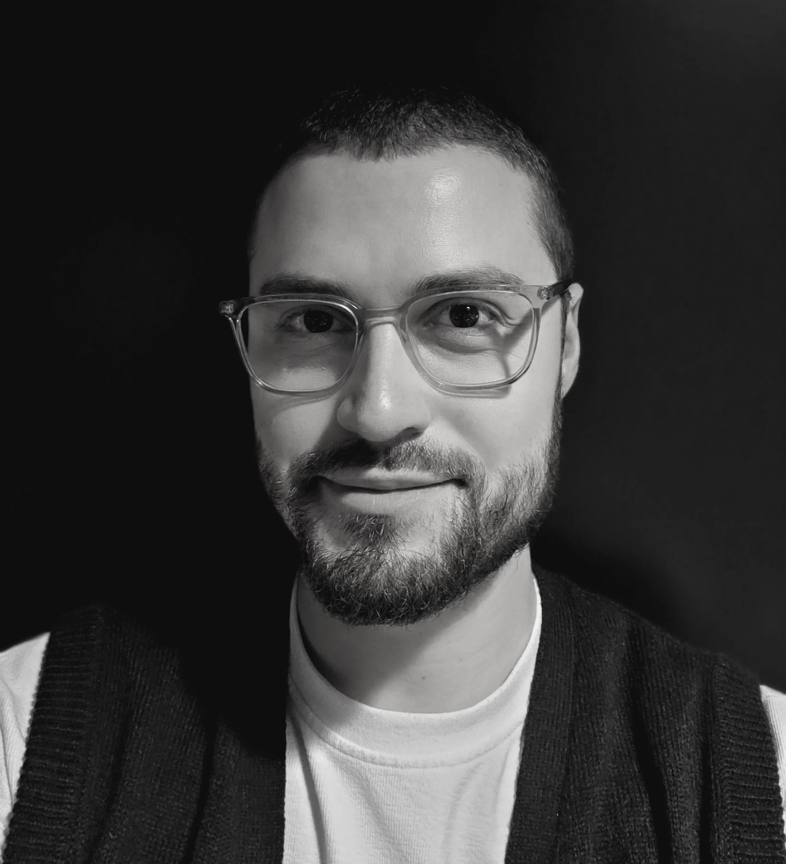 Black and white photo of a man with short hair, glasses, a beard, wearing a white shirt and a dark vest, smiling slightly, against a plain dark background.