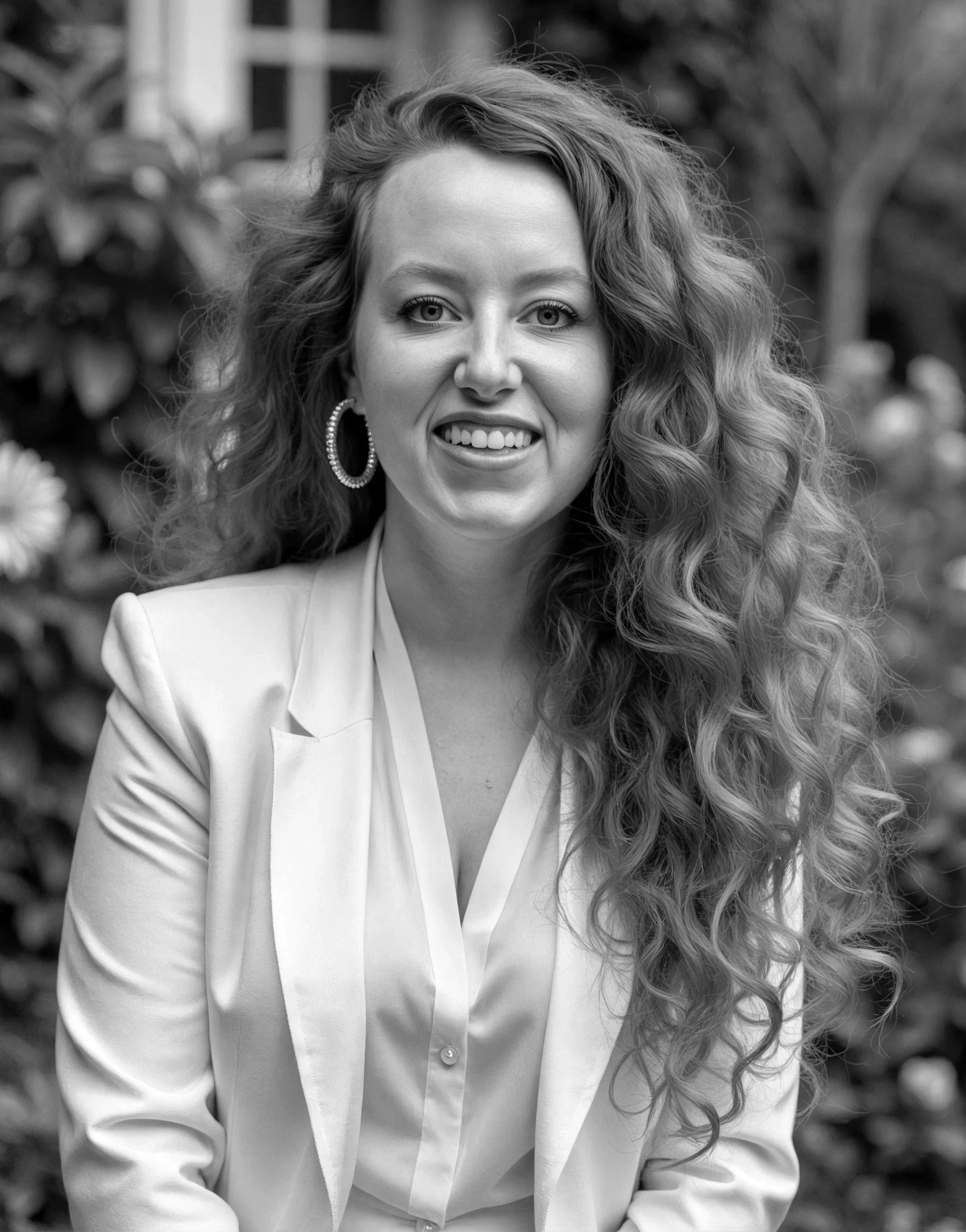A woman with long, curly hair smiling in black and white, wearing a white blazer and hoop earrings, outdoors with blurred plants in the background.