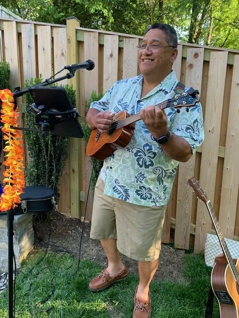 Man playing ukulele outdoors in a backyard with a wooden fence, wearing a Hawaiian shirt and shorts, smiling.