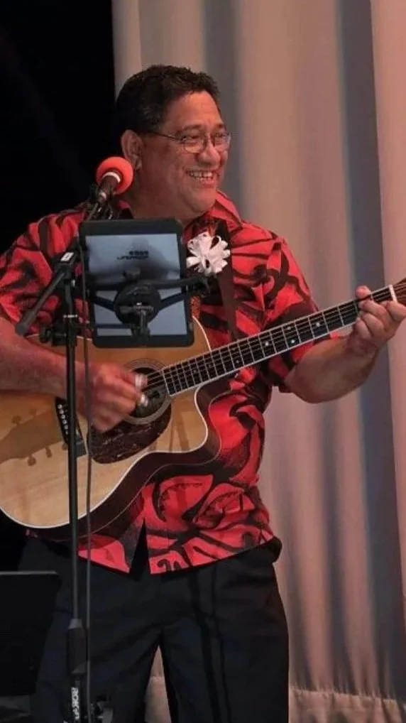 A man wearing a red Hawaiian shirt with black patterns, glasses, and a white flower clip on his shirt, is playing an acoustic guitar and smiling on stage with a microphone in front of him.