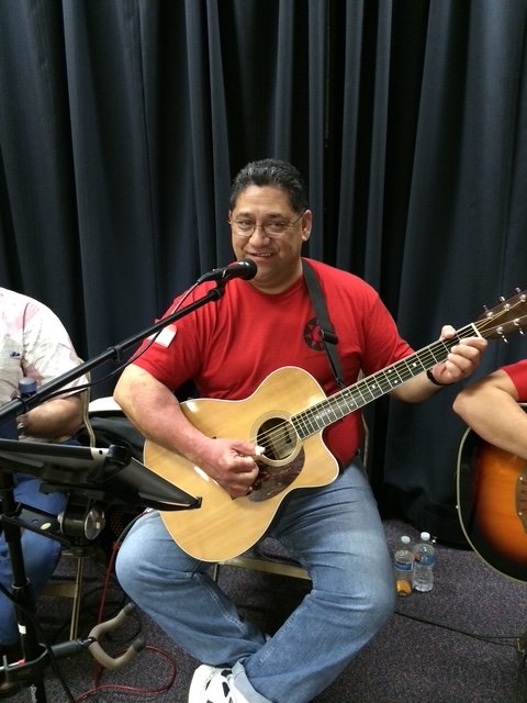 A man in a red T-shirt playing an acoustic guitar while sitting on a chair with a microphone in front of him, in front of a dark curtain backdrop.