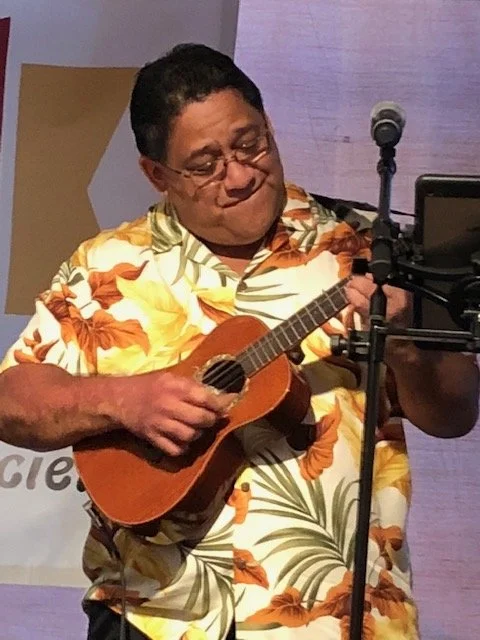 Man playing ukulele on stage, wearing a tropical floral shirt with a microphone nearby.
