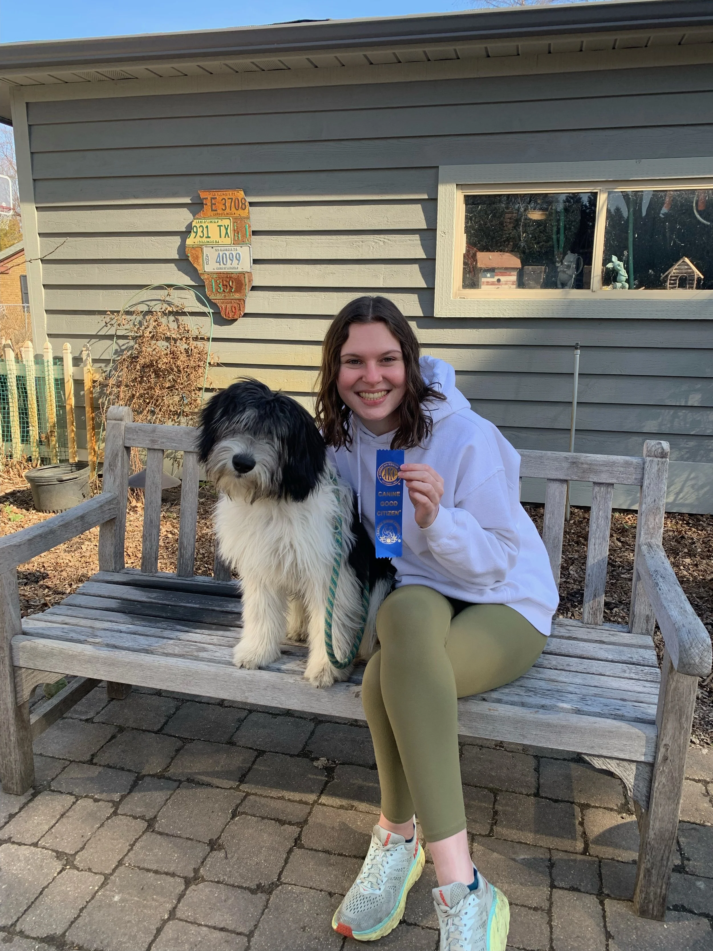 A woman sitting on a bench with her black and white fluffy dog. The woman holds up a blue ribbon that they won