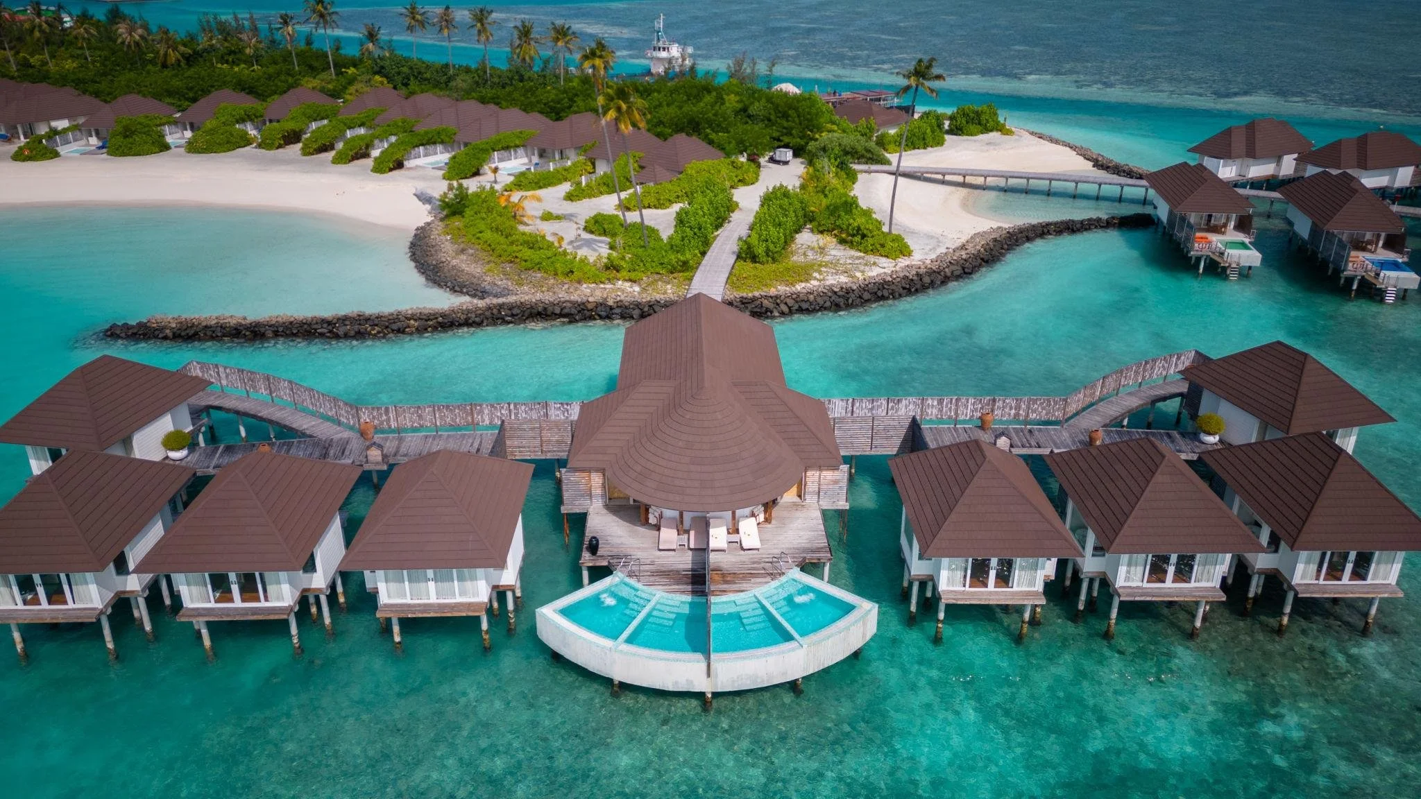 Overwater bungalows with brown roofs over turquoise water, connecting to a central building with an infinity pool, with a sandy beach, palm trees, and a lush green island in the background.