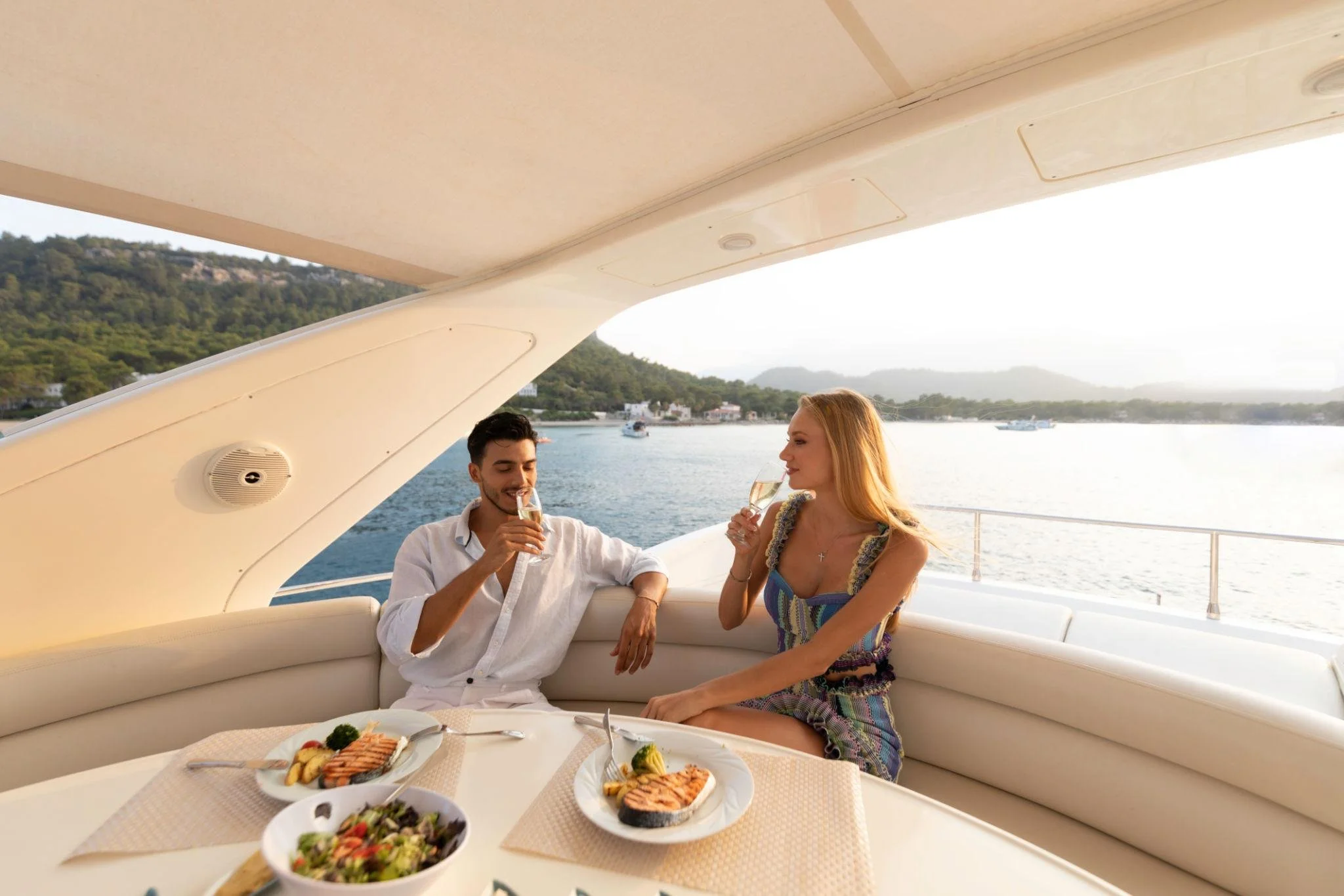 A man and woman enjoying drinks at a dining table on a yacht with a water and mountainous landscape in the background.