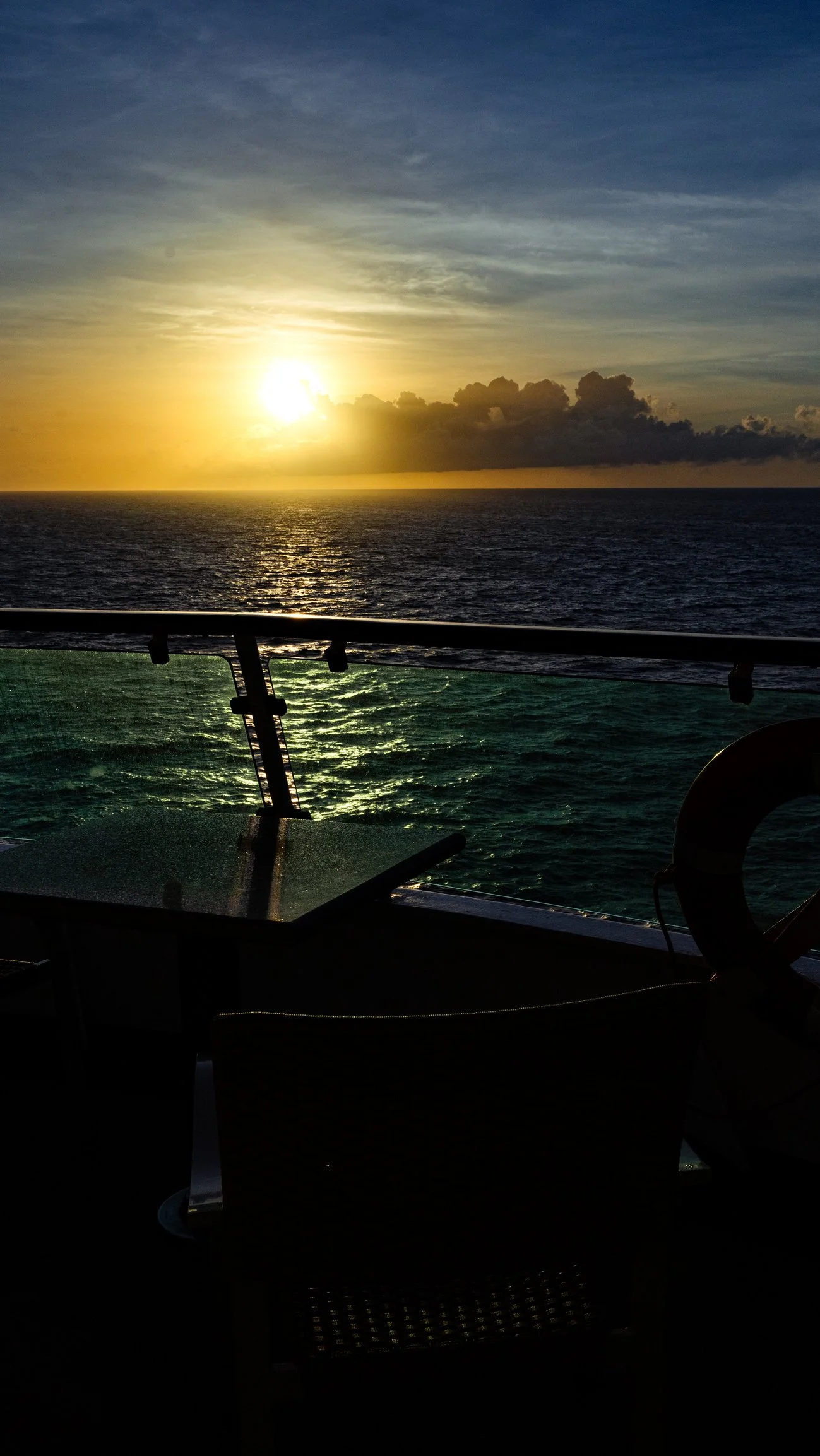 Sunset view over the ocean from a boat deck, with clouds in the sky and water reflecting the sunlight.