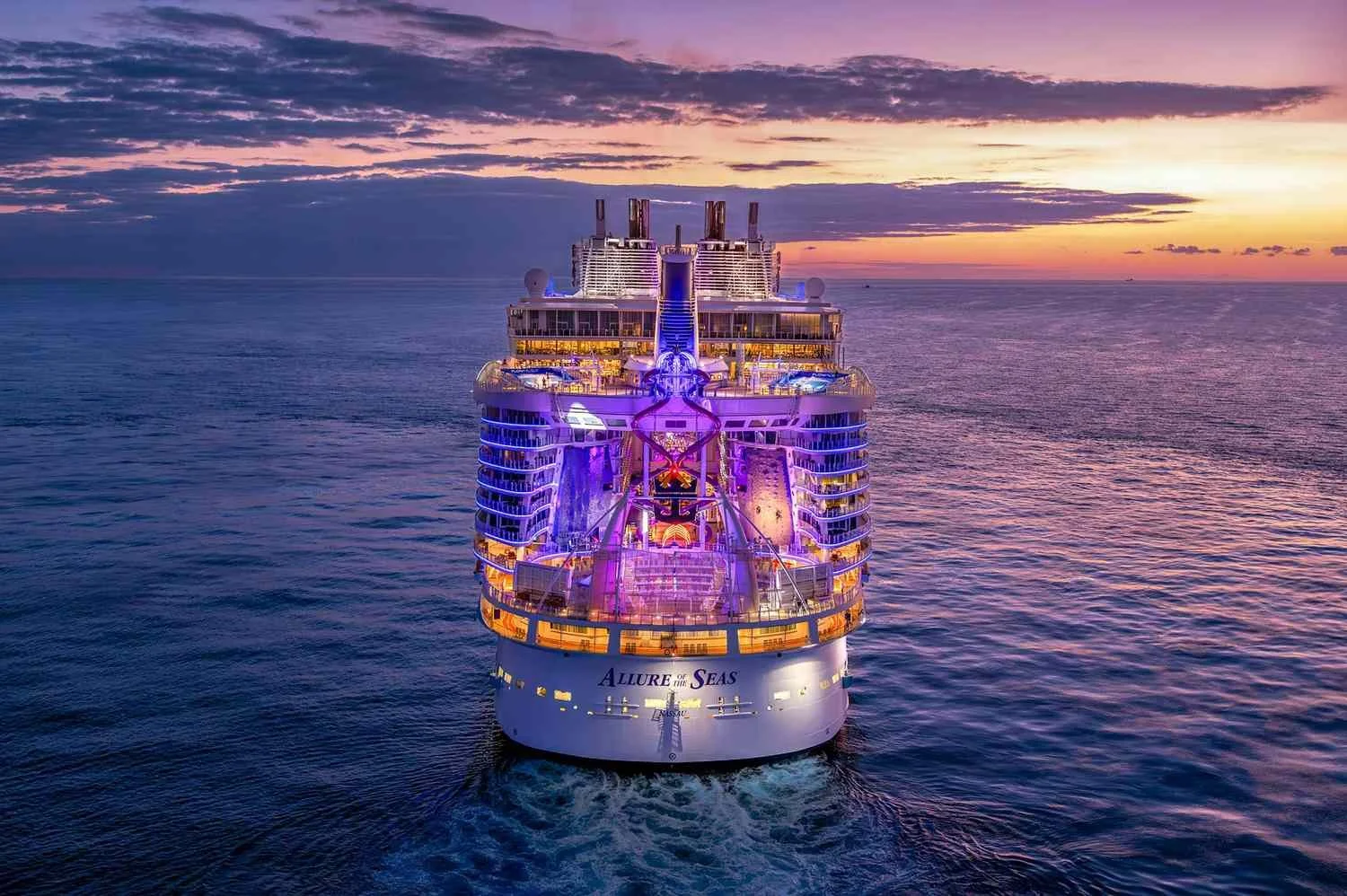 A large luxury cruise ship named Allure of the Seas sailing on the ocean at sunset, illuminated with colorful lights.