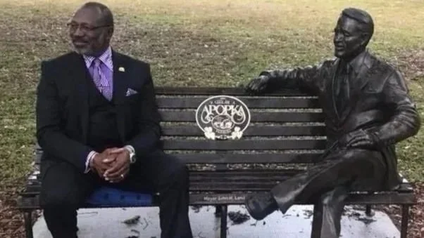 A man in a black suit with glasses sitting on a park bench, next to a bronze statue of a man seated on the bench, with the statue's arm resting on the back of the bench.