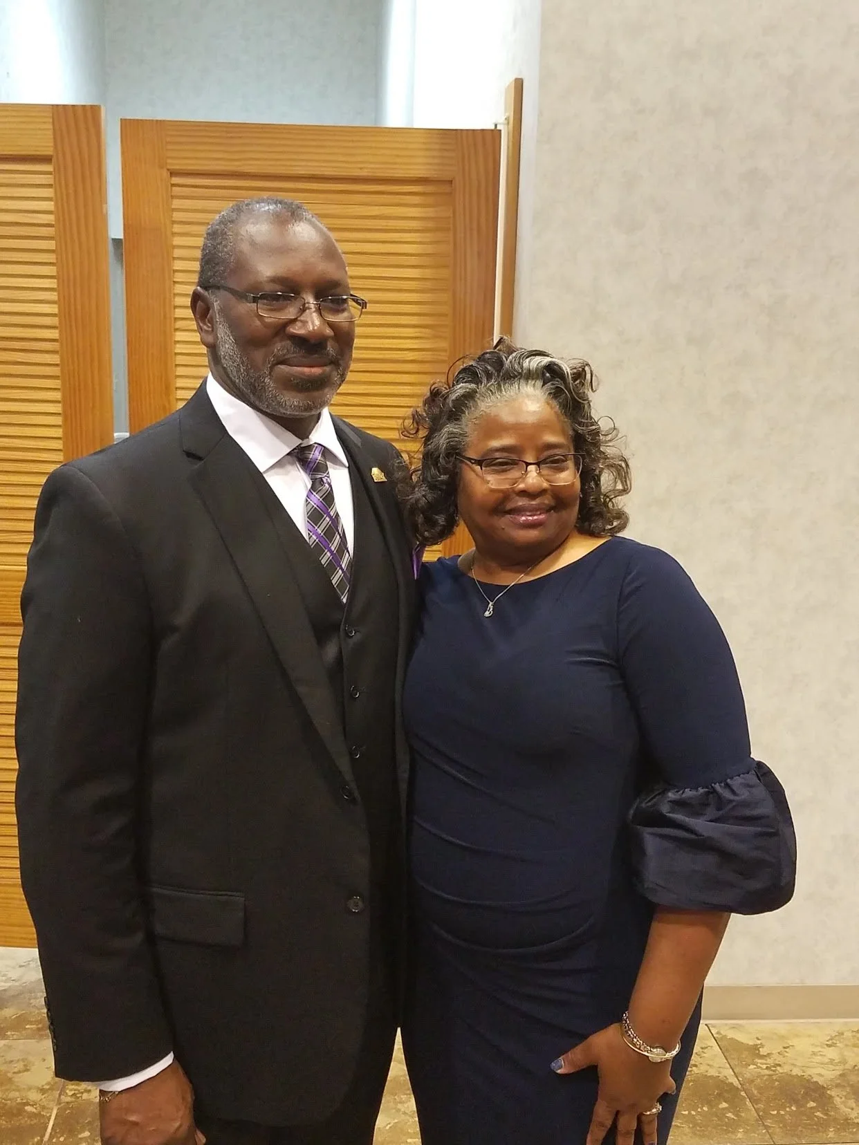 A man and woman dressed in formal attire standing together in an indoor setting, smiling at the camera.