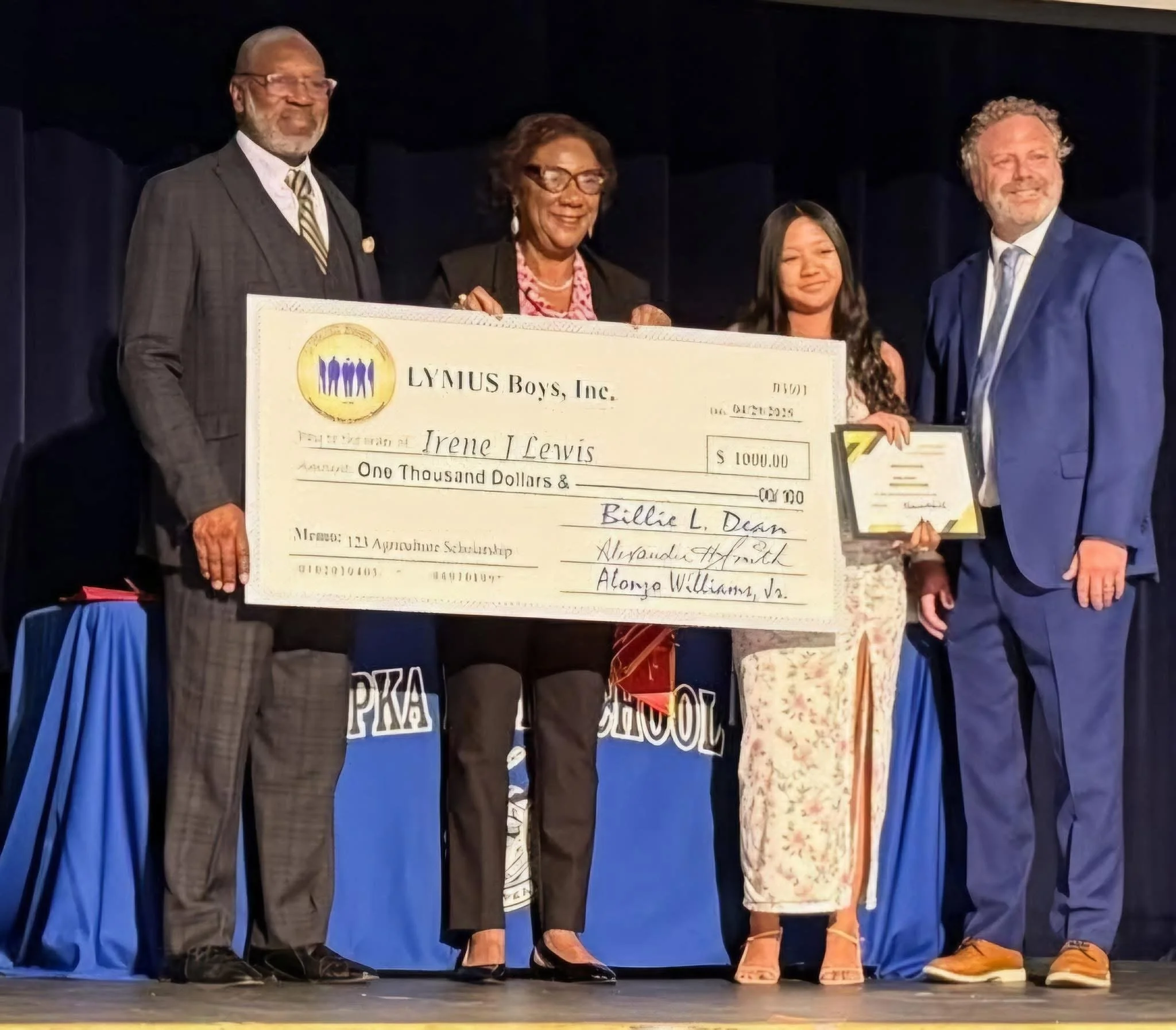People on stage holding an oversized check and a framed certificate during an award ceremony, with a blue backdrop and table draped in blue.