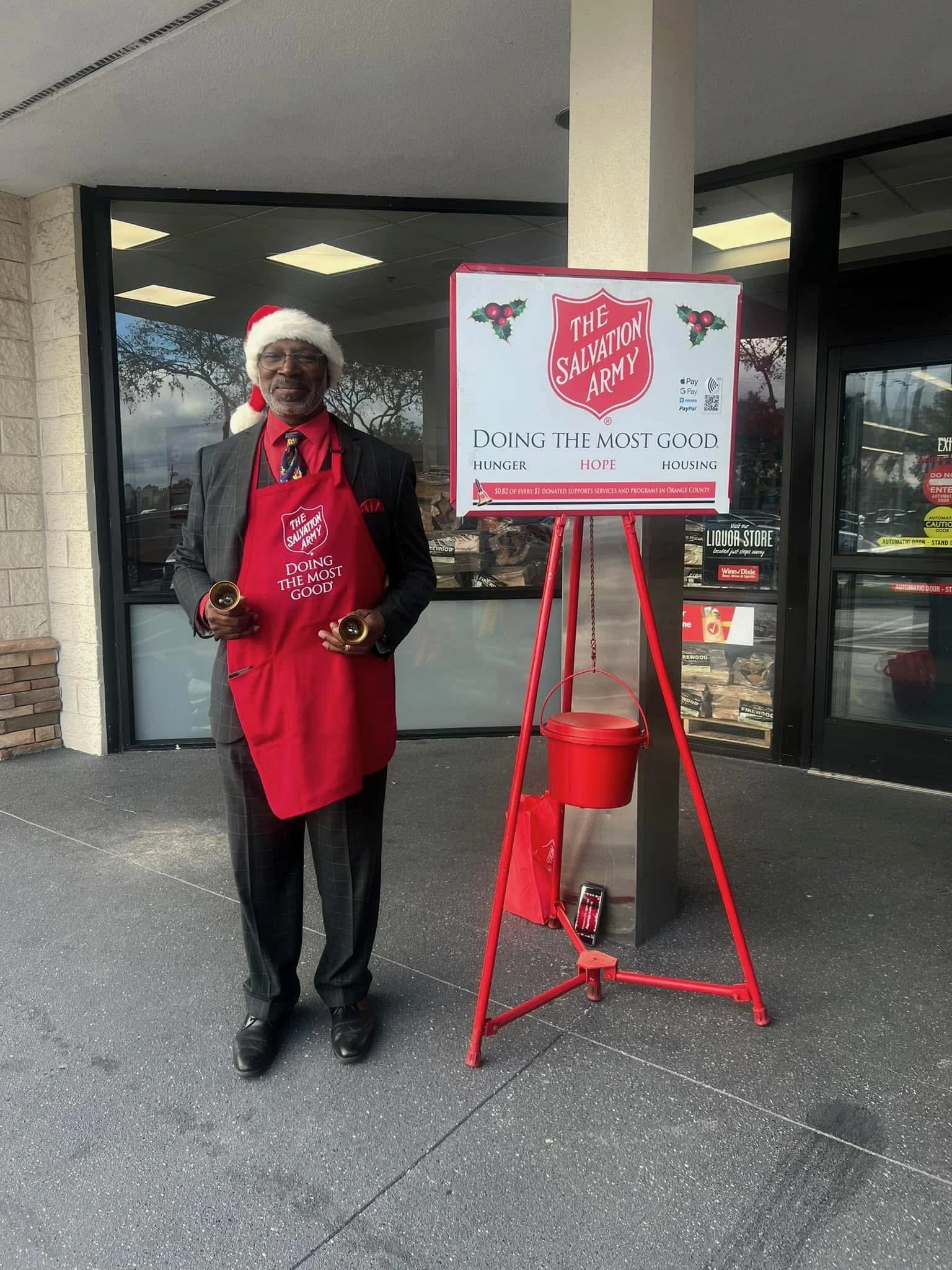 Man wearing a Santa hat and an apron with Salvation Army logo, standing outside a store next to a red donation kettle and a sign for The Salvation Army with Christmas decorations.