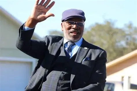 A man dressed in a suit, waving and smiling outdoors on a sunny day, wearing sunglasses and a flat cap.