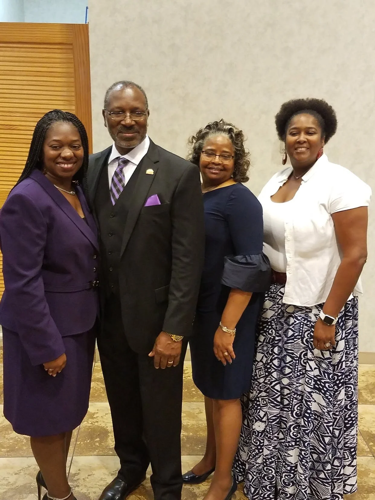 Group of four women and one man posing together indoors, dressed in formal and professional attire, smiling at the camera.