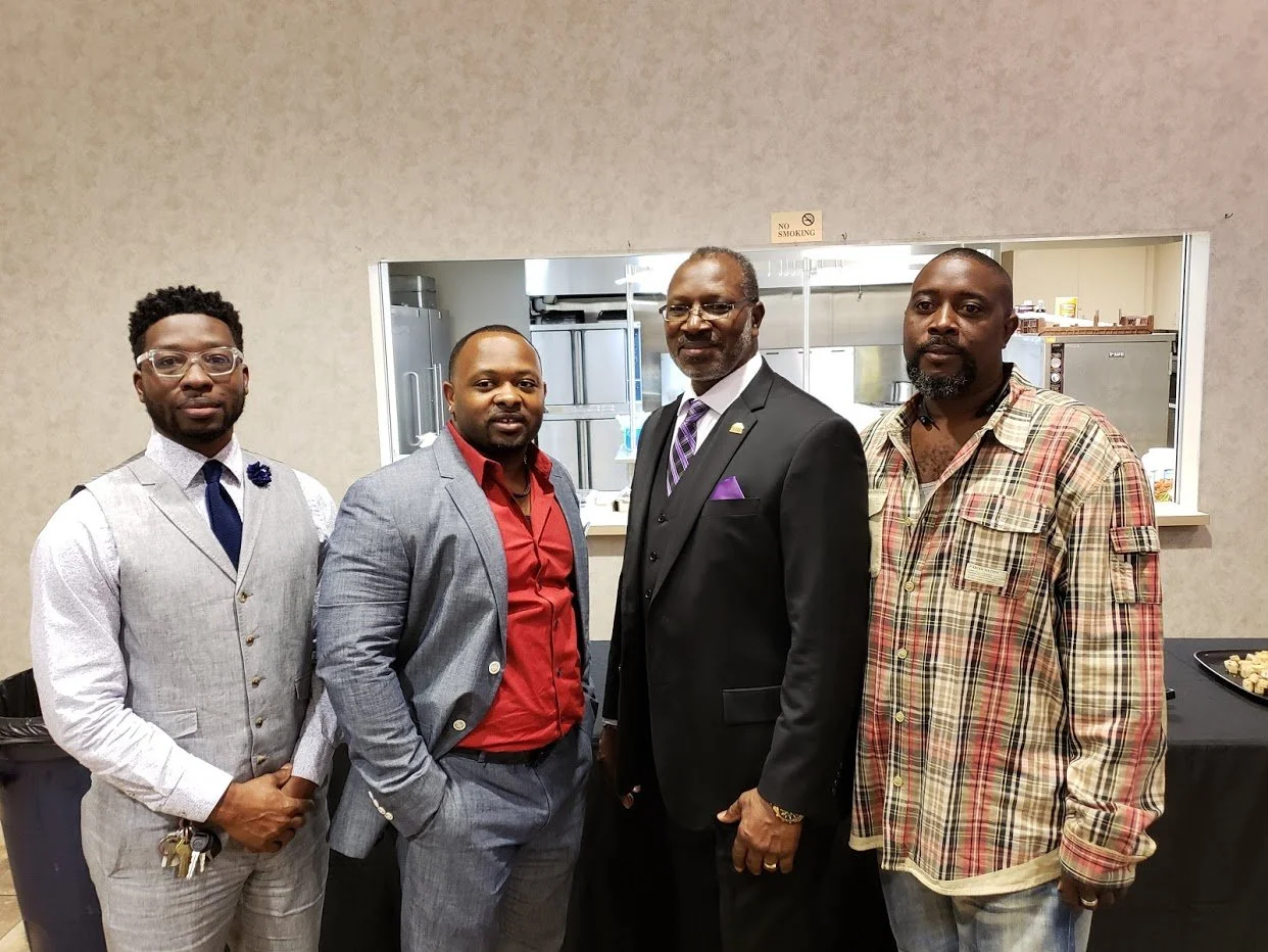 Four men posing together indoors in front of a wall with a window opening to a kitchen area.