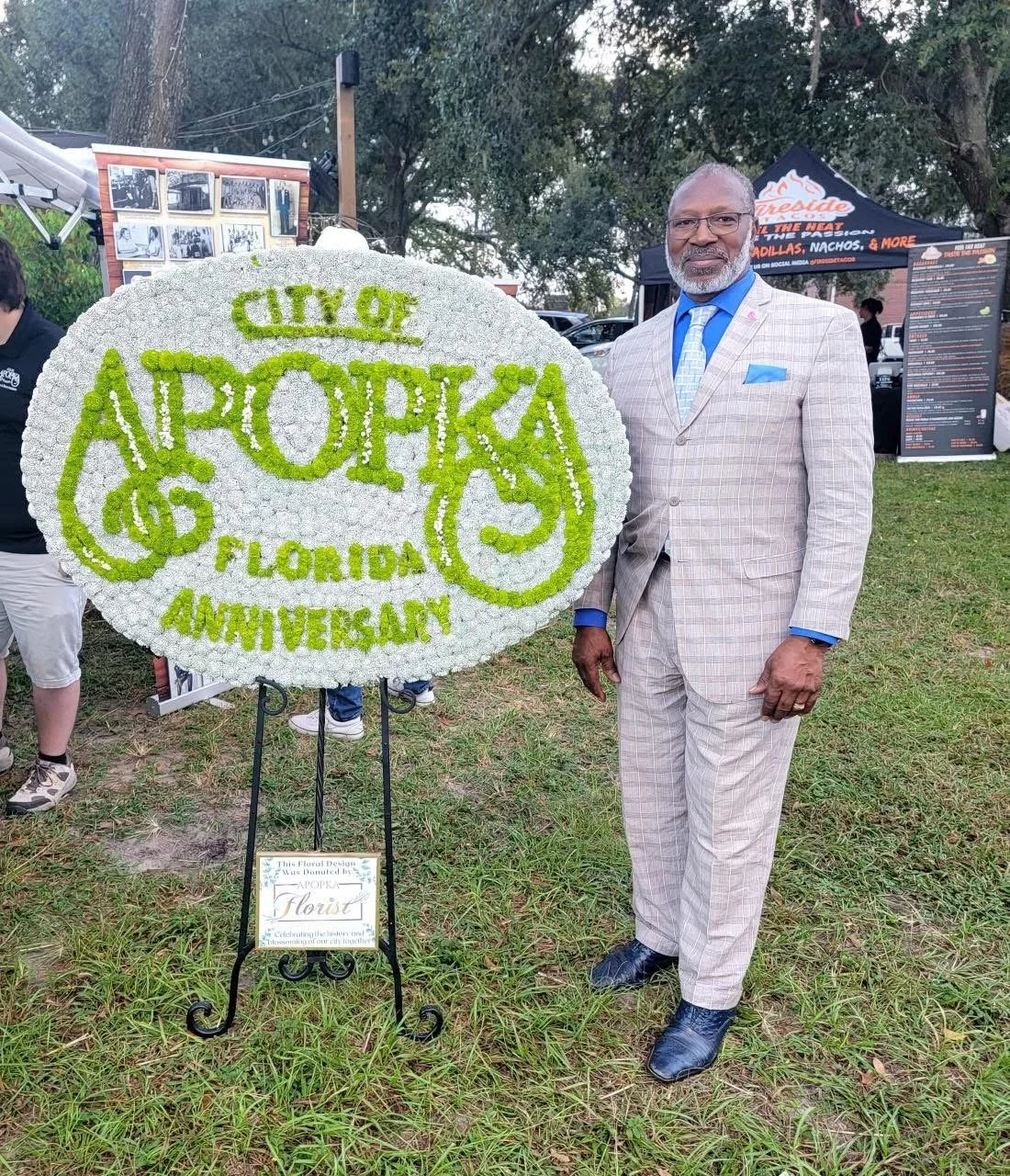 A man in a checked suit and blue tie standing next to a floral display with a sign celebrating the City of Apopka, Florida's anniversary.