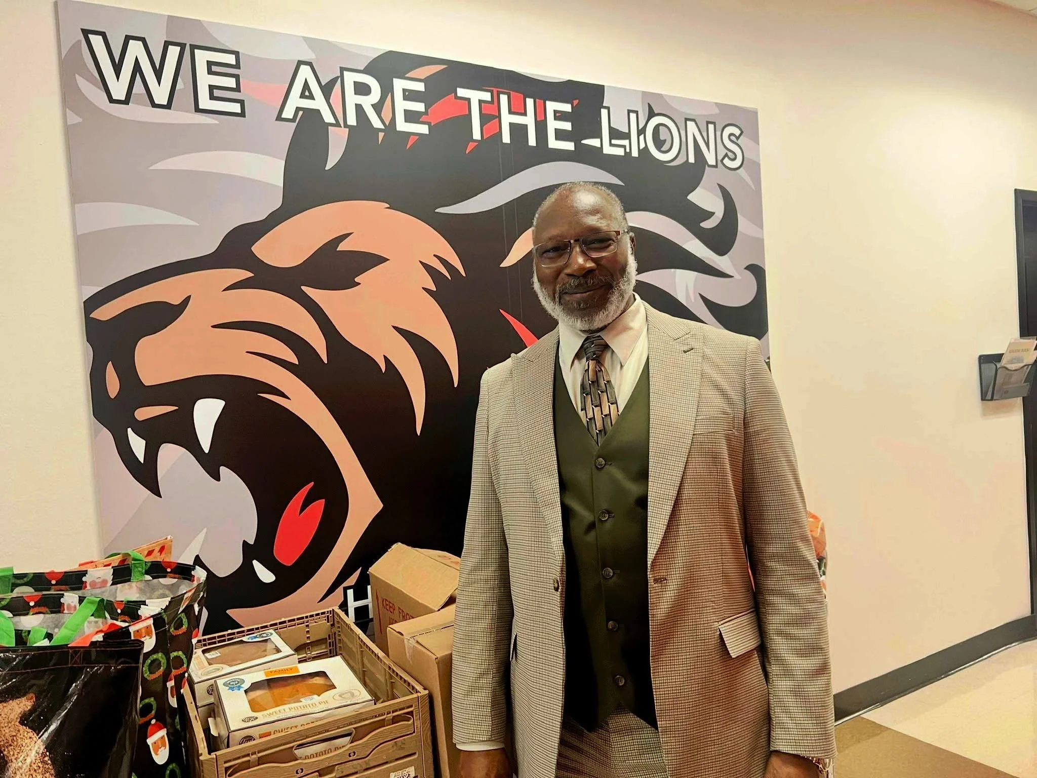 A man with glasses, gray beard, wearing a beige checked suit, standing in front of a mural with a roaring lion and the words "WE ARE THE LIONS."