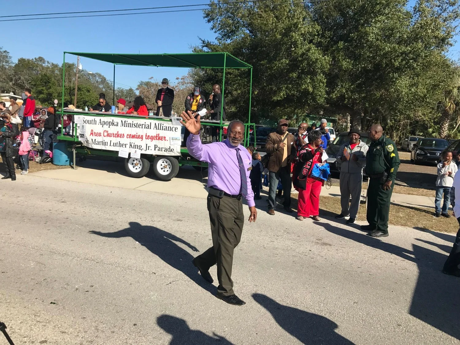 Man in purple shirt waving at parade, with marching group and float in background, celebrating Martin Luther King Jr. Day