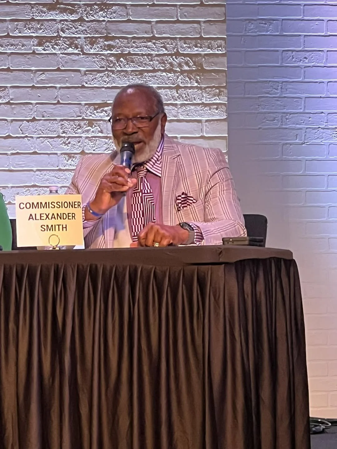 An elderly man with glasses and a beard, wearing a light pink striped suit and a patterned shirt, speaking into a microphone at a formal event. A nameplate in front of him reads 'Commissioner Alexander Smith'. The background features a brick wall ill