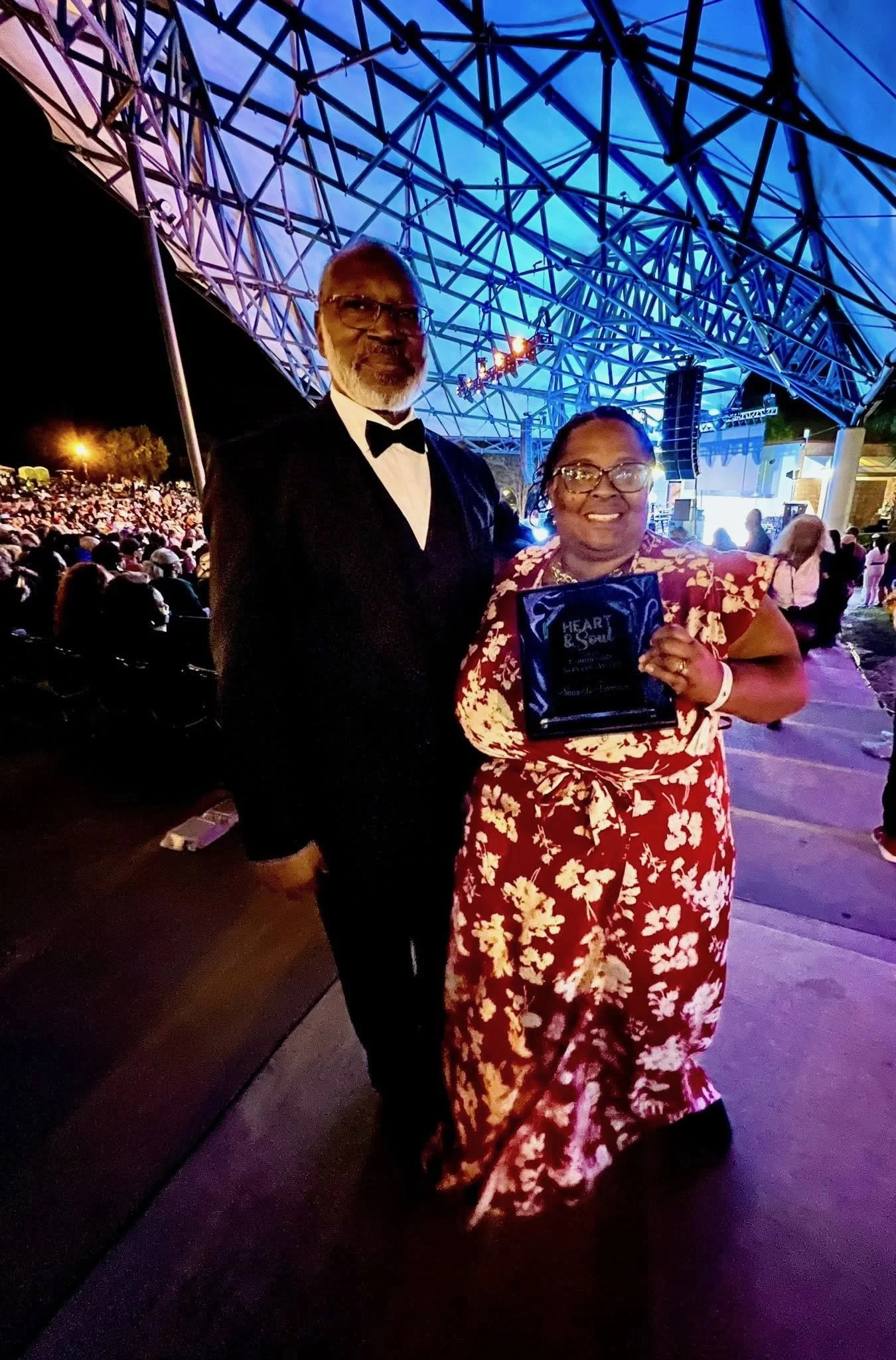 A man and a woman at an outdoor event during nighttime. The man is dressed in a tuxedo, and the woman is wearing a red floral dress, holding a plaque or award. There is a large crowd and a stage with lighting in the background.