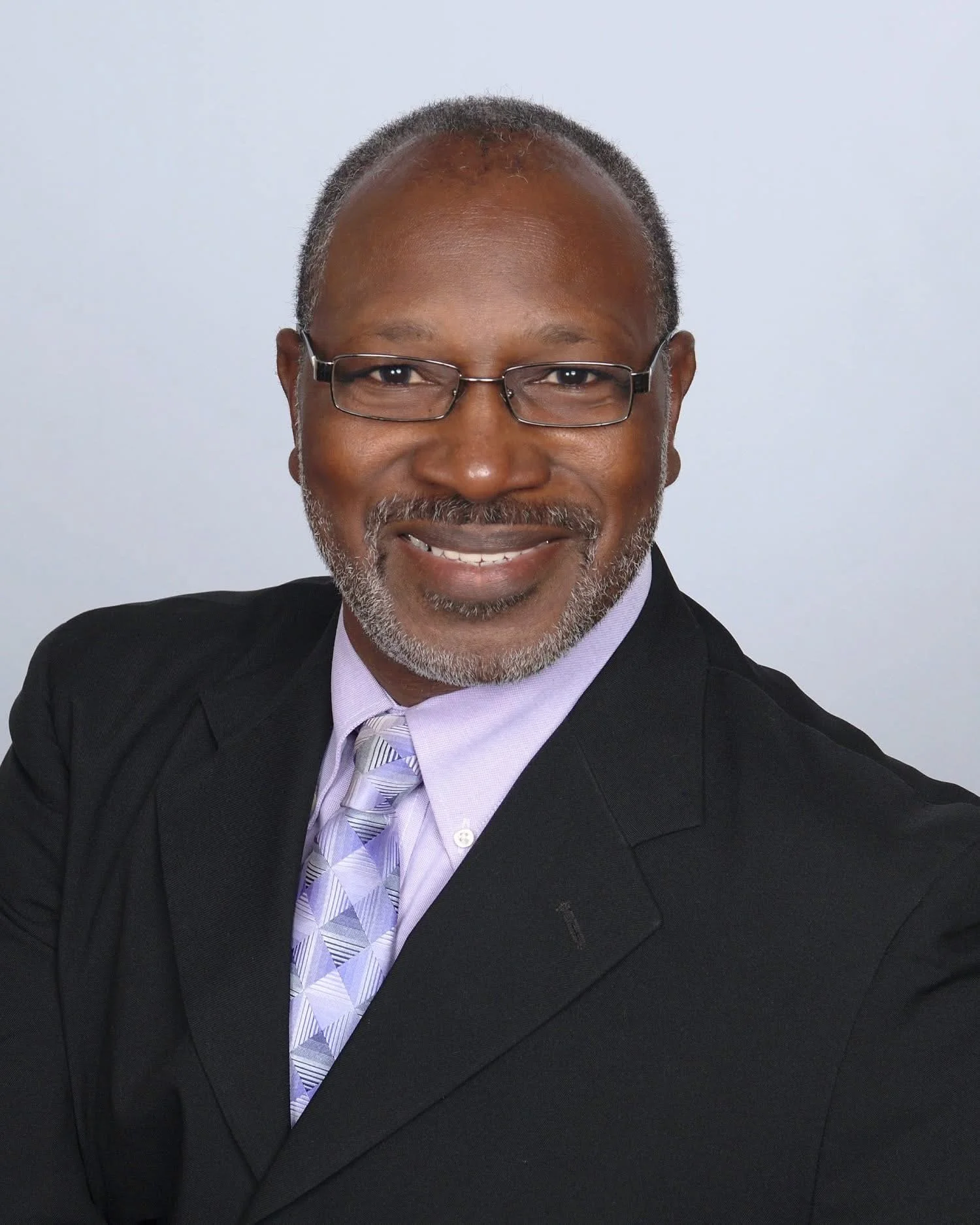 Portrait of a middle-aged African American man wearing glasses, a black suit, a light purple shirt, and a patterned purple and white tie, smiling against a plain light background.