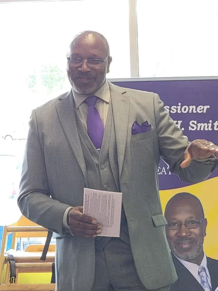 A man in a light gray suit, purple tie, and glasses standing indoors, holding papers in one hand and gesturing with the other, with a poster of himself behind him.