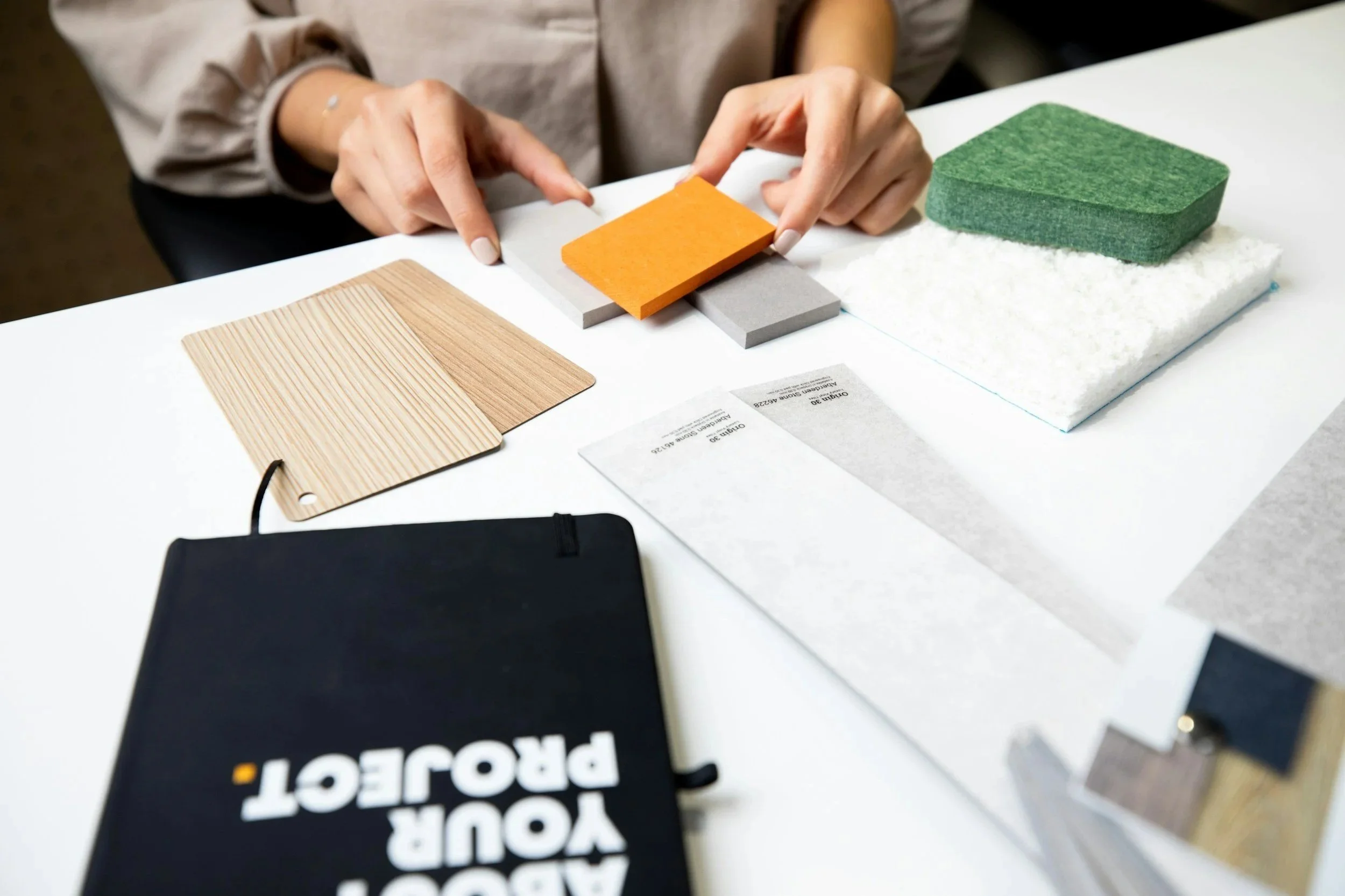 Person arranging small sample tiles of wood, fabric, and color options on a white table, with a black notebook labeled "Your Project" nearby.