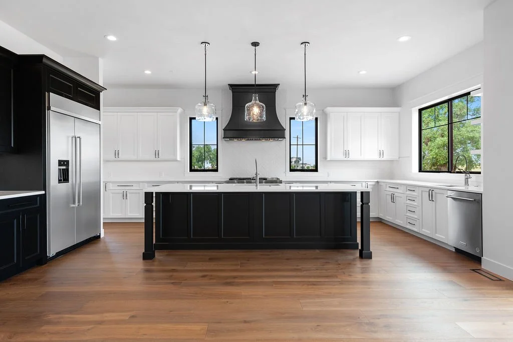 Modern kitchen with black and white cabinetry, wooden flooring, large windows, and pendant lights over the island.