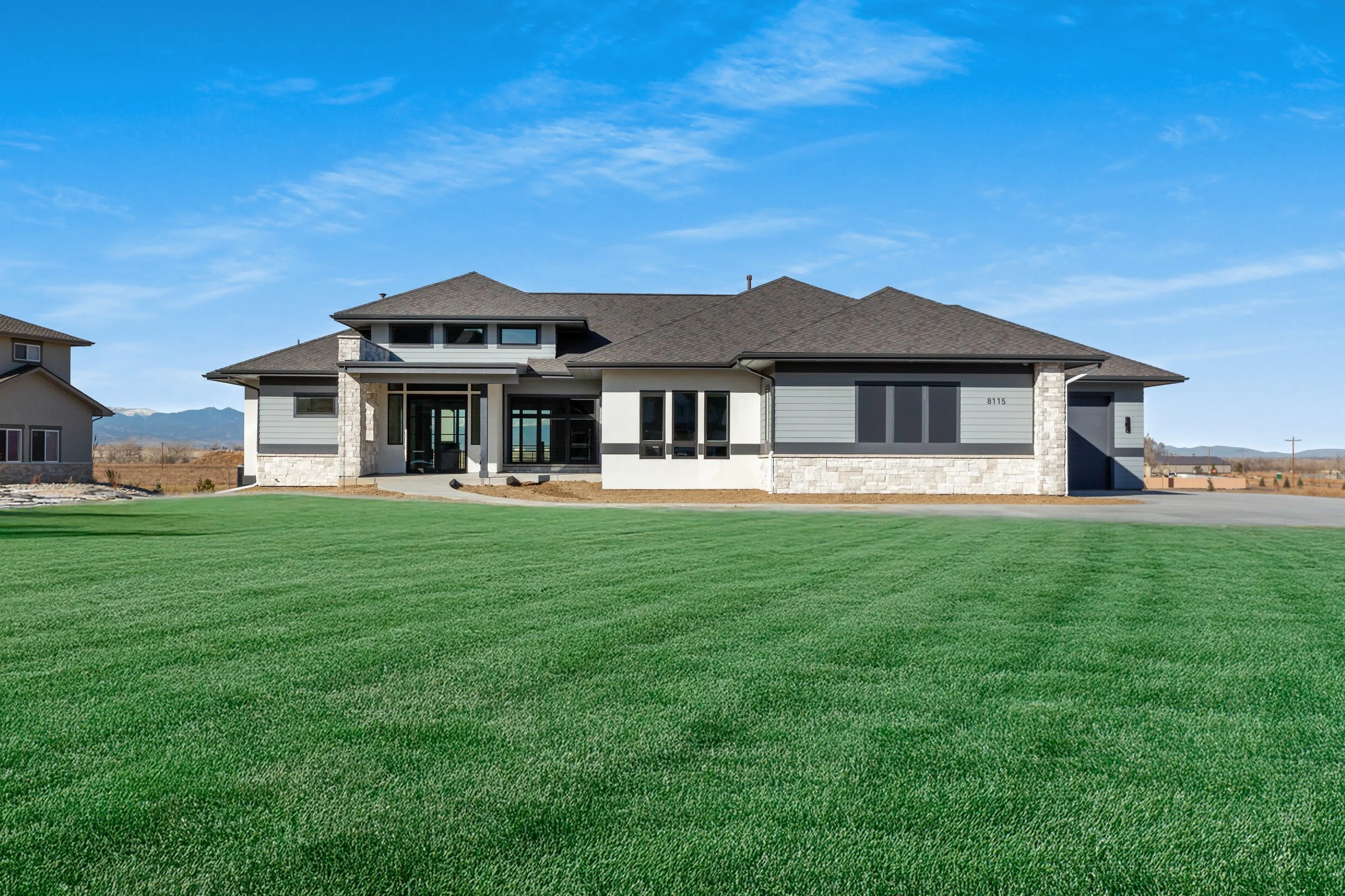 Modern house with a gabled roof, white and gray exterior, and large windows, surrounded by a well-maintained green lawn under a clear blue sky.