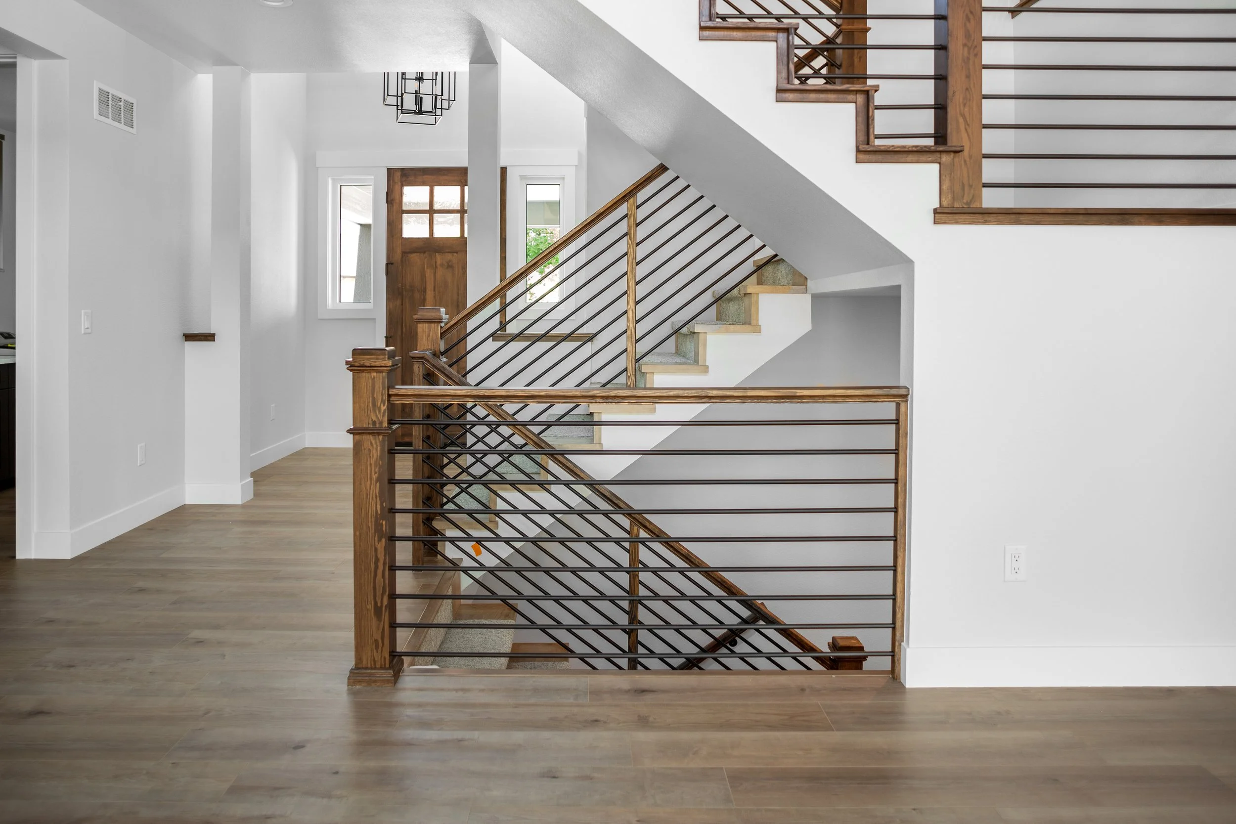 Interior view of a modern house with a staircase featuring wood handrails and black metal balusters, white walls, light hardwood floors, and a wooden door with glass panels in the background.