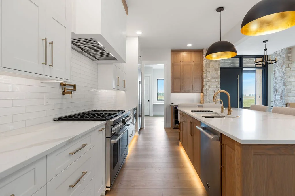 Modern kitchen with white and wood cabinetry, black hanging pendant lights, a large island with a white countertop and brass fixtures, stainless steel appliances, a stone accent wall, and sliding glass doors leading to an outdoor view.