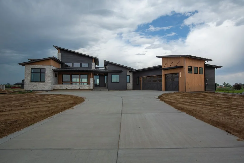 Modern house with concrete driveway, surrounded by dirt, under a cloudy sky.